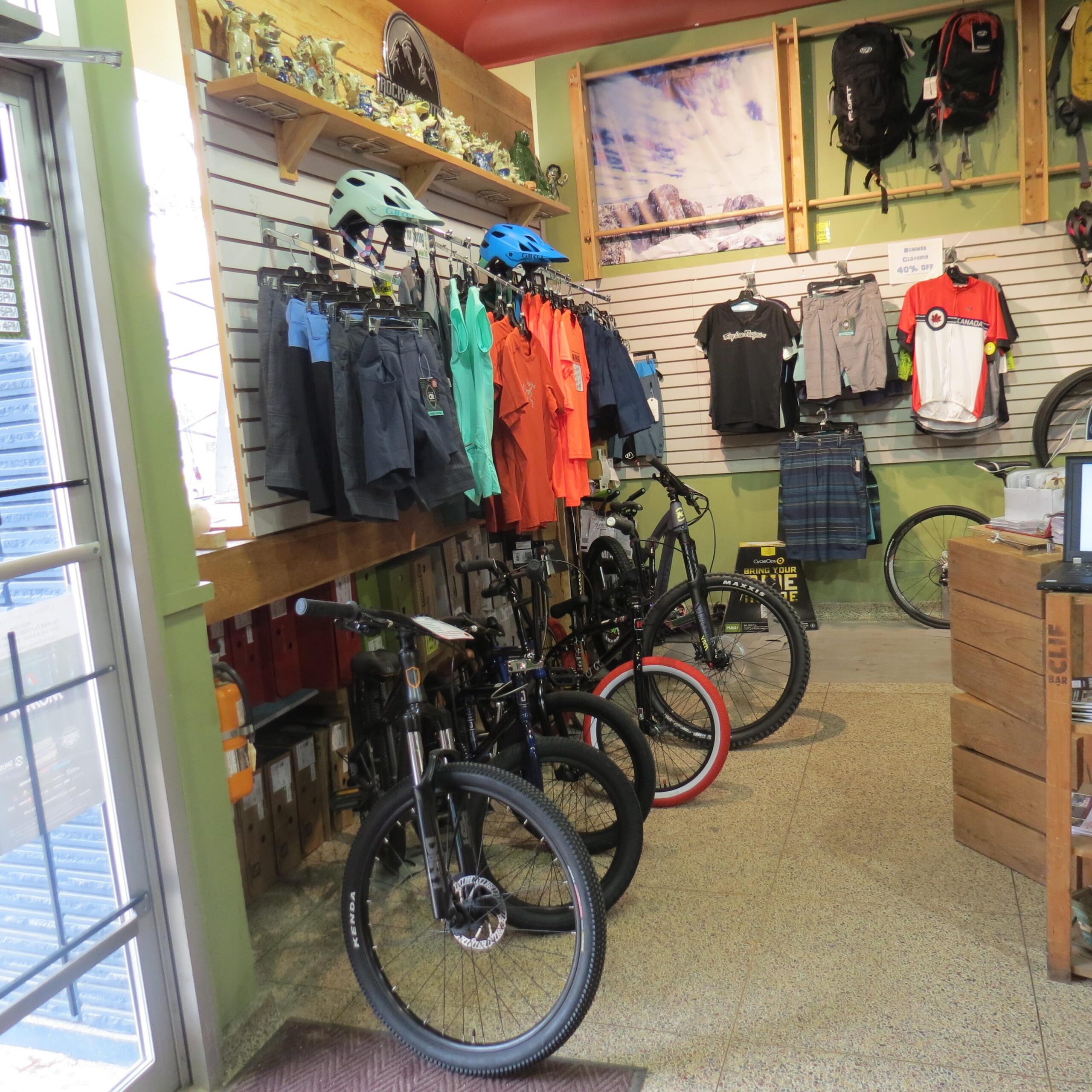 Interior of a bike shop featuring several mountain bikes lined up on the floor. On the walls, there are shelves displaying helmets and various clothing items, including shorts and t-shirts in vibrant colors. Other biking gear, such as backpacks, is also visible. The space has a welcoming atmosphere with natural light coming in from a window.