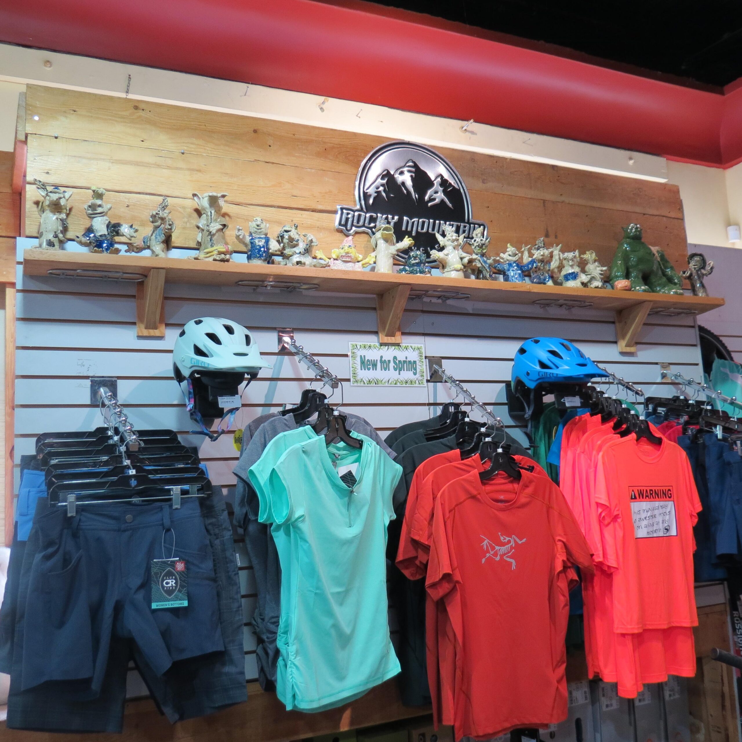 A display of outdoor clothing and accessories featuring a variety of colorful shirts, shorts, and helmets. The items are arranged neatly on hangers, with a wooden shelf above displaying decorative figurines. A sign labeled "New for Spring" is prominently featured, along with a logo for "Rocky Mountain" at the top.