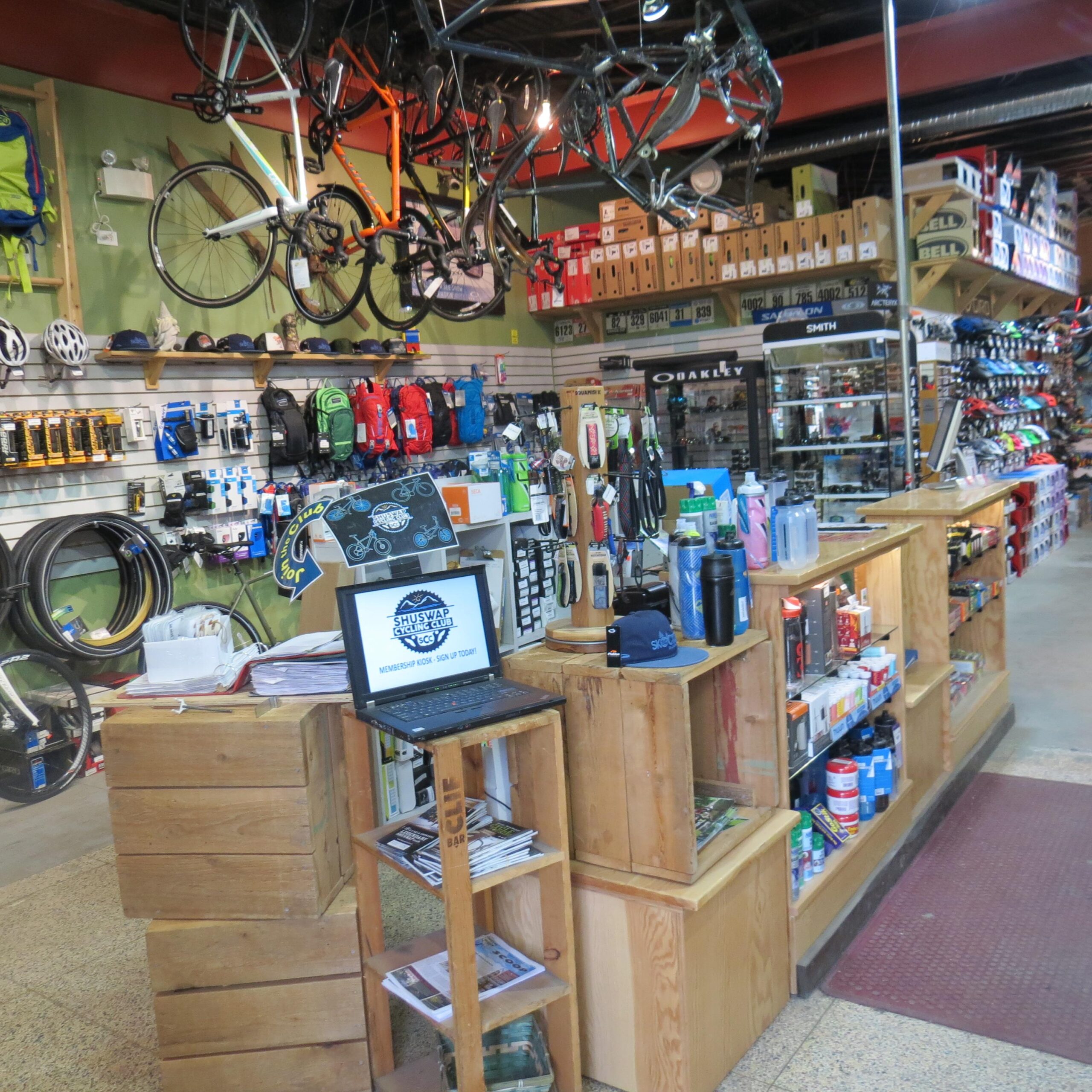 A brightly lit bike shop interior featuring a wooden sales counter with a laptop displaying promotional content. Bikes hang from the ceiling above shelves stocked with various cycling gear, including helmets, backpacks, and water bottles. The walls are lined with colorful accessories, demonstrating a vibrant and organized retail space focused on cycling products.