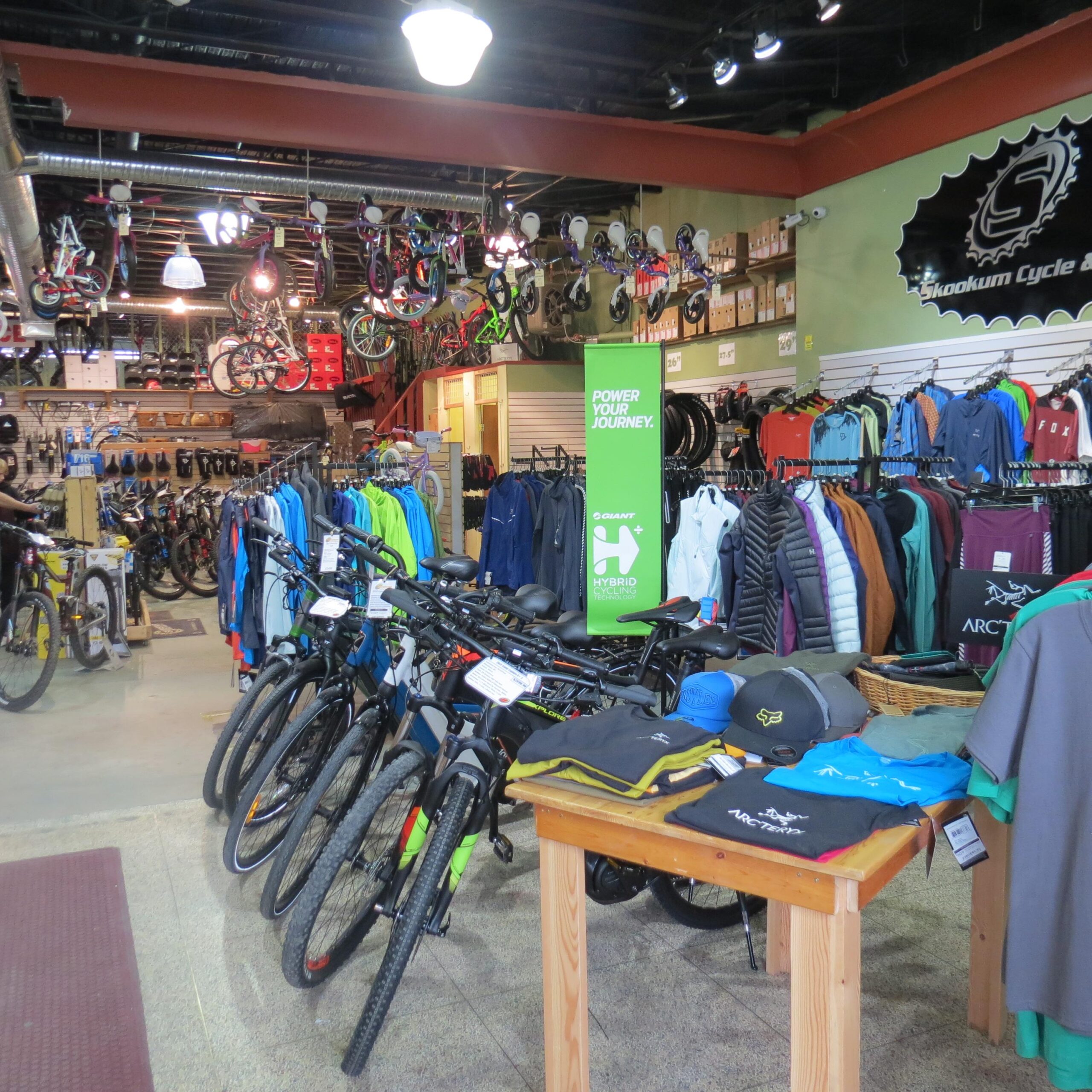 A bicycle shop interior featuring a variety of bicycles displayed prominently. The foreground includes several mountain bikes lined up, while colorful apparel and accessories hang on the walls. A green promotional sign reads "Power Your Journey." The shop has a well-organized layout, with helmets and cycling gear also visible.