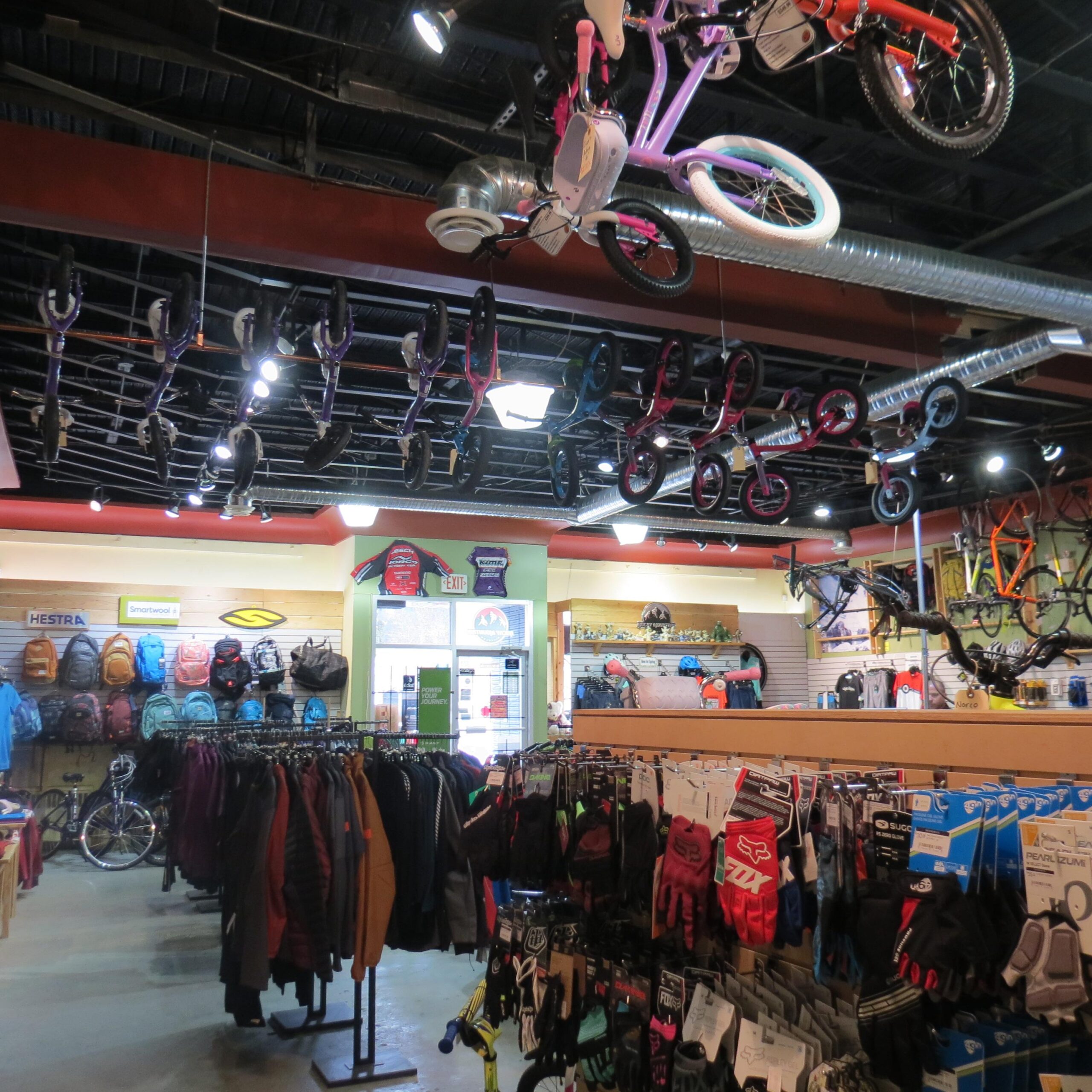 A colorful retail environment featuring various bicycles hanging from the ceiling. The store showcases a wide range of clothing and accessories, with racks filled with jackets and gloves in the foreground, while backpacks and helmets are displayed on the walls. Bright lighting illuminates the space, creating an inviting atmosphere for customers.