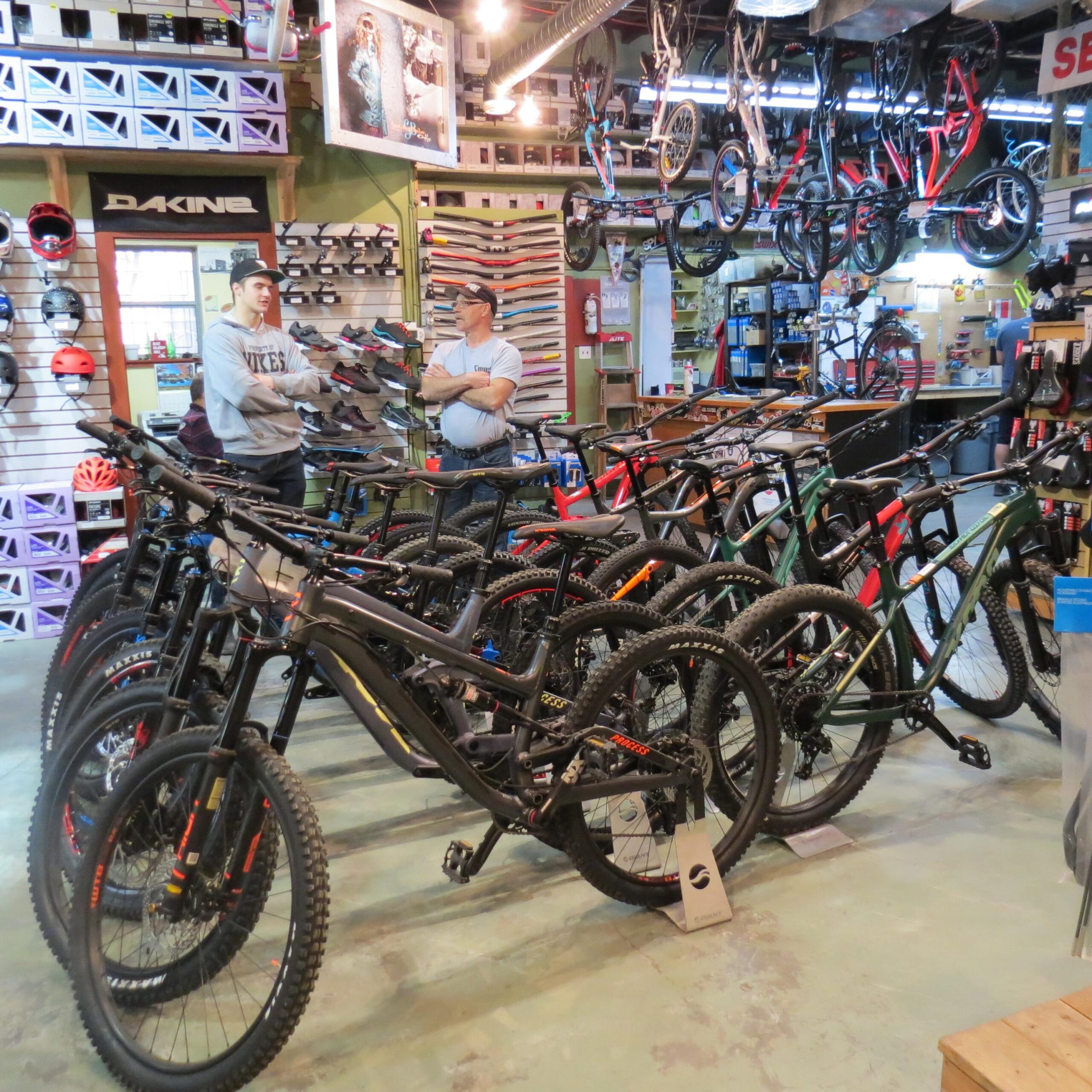 Two men are engaged in conversation in a well-stocked bike shop featuring a wide range of mountain bikes. Multiple bikes of various colors are lined up in the foreground, while helmets and bike accessories are displayed on the walls behind them. The shop has a vibrant, active atmosphere with equipment for cycling enthusiasts.