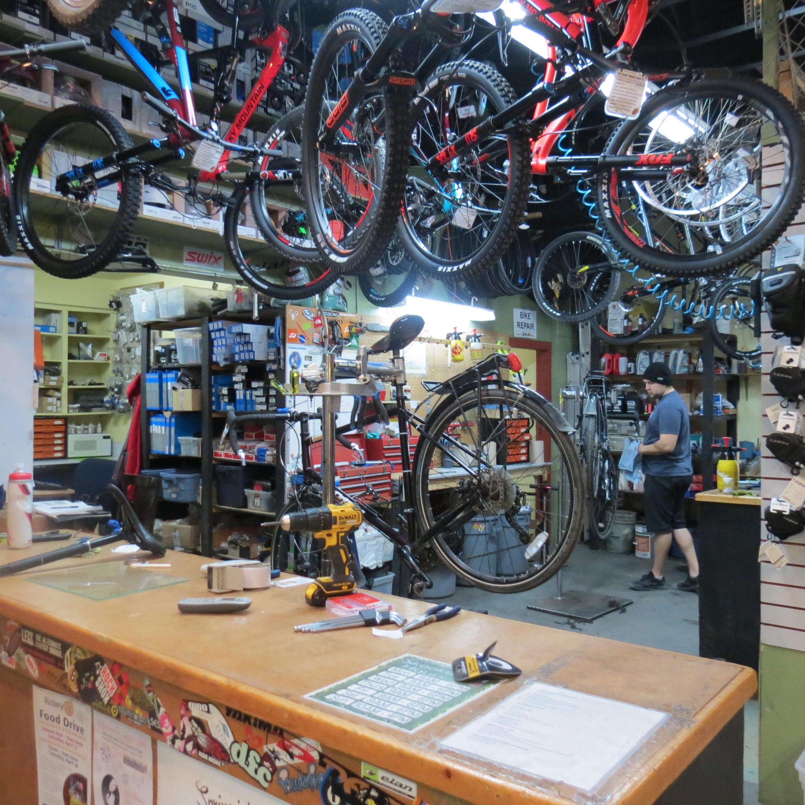 Alt text: Inside a bike shop, displaying a wooden counter with tools and a service menu. Various bicycles hang from the ceiling, and shelves filled with bike parts and accessories are visible in the background. A person is seen working on a bike in the workshop area.