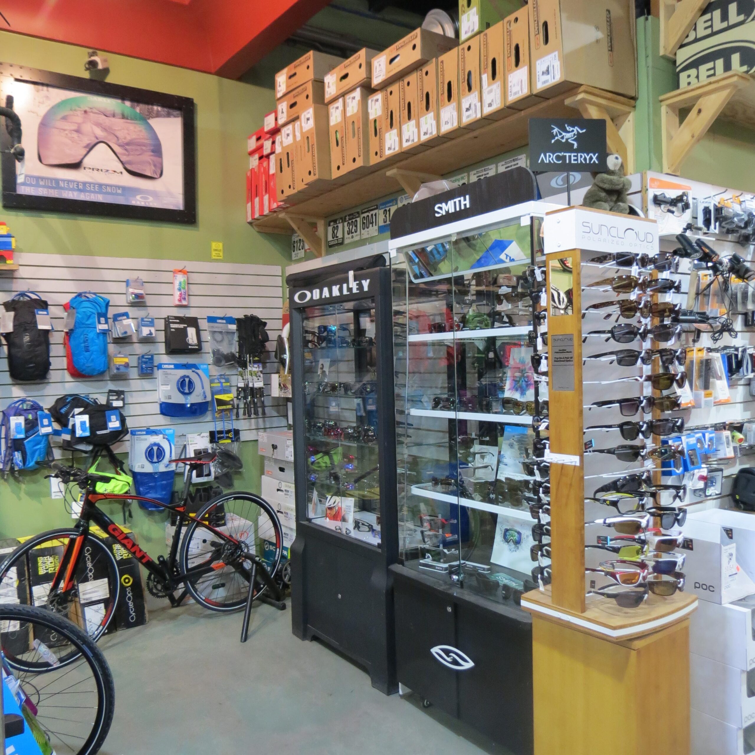 Interior of a sporting goods store featuring a display of sunglasses from brands like Oakley and Smith. The background includes backpacks, cycling gear, and various sporting equipment. A bicycle is positioned in the foreground, and shelves stocked with boxed items are visible overhead.