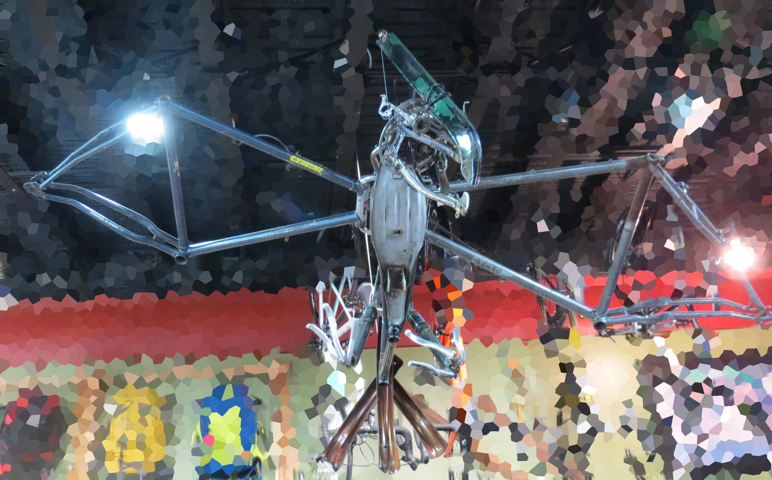 A mechanical sculpture resembling a bird, made from various metal parts, including bicycle frames and other components. The artwork is suspended from the ceiling and features glowing lights, set against a colorful, abstract background of hanging backpacks and wall decor.