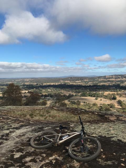 Kona Honzo: A mountain bike resting on rocky terrain overlooking a picturesque landscape with rolling hills under a partly cloudy sky.
