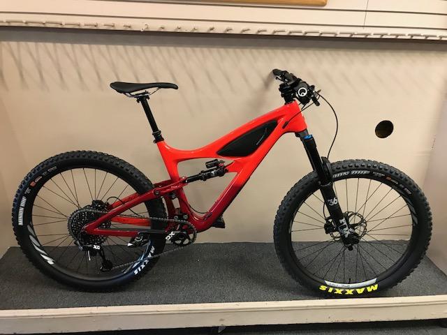 Ibis Mojo HD4: A red mountain bike displayed on a stand, featuring a sleek design, full suspension, wide tires, and a modern drivetrain. The background includes a simple surface with a beige wall.