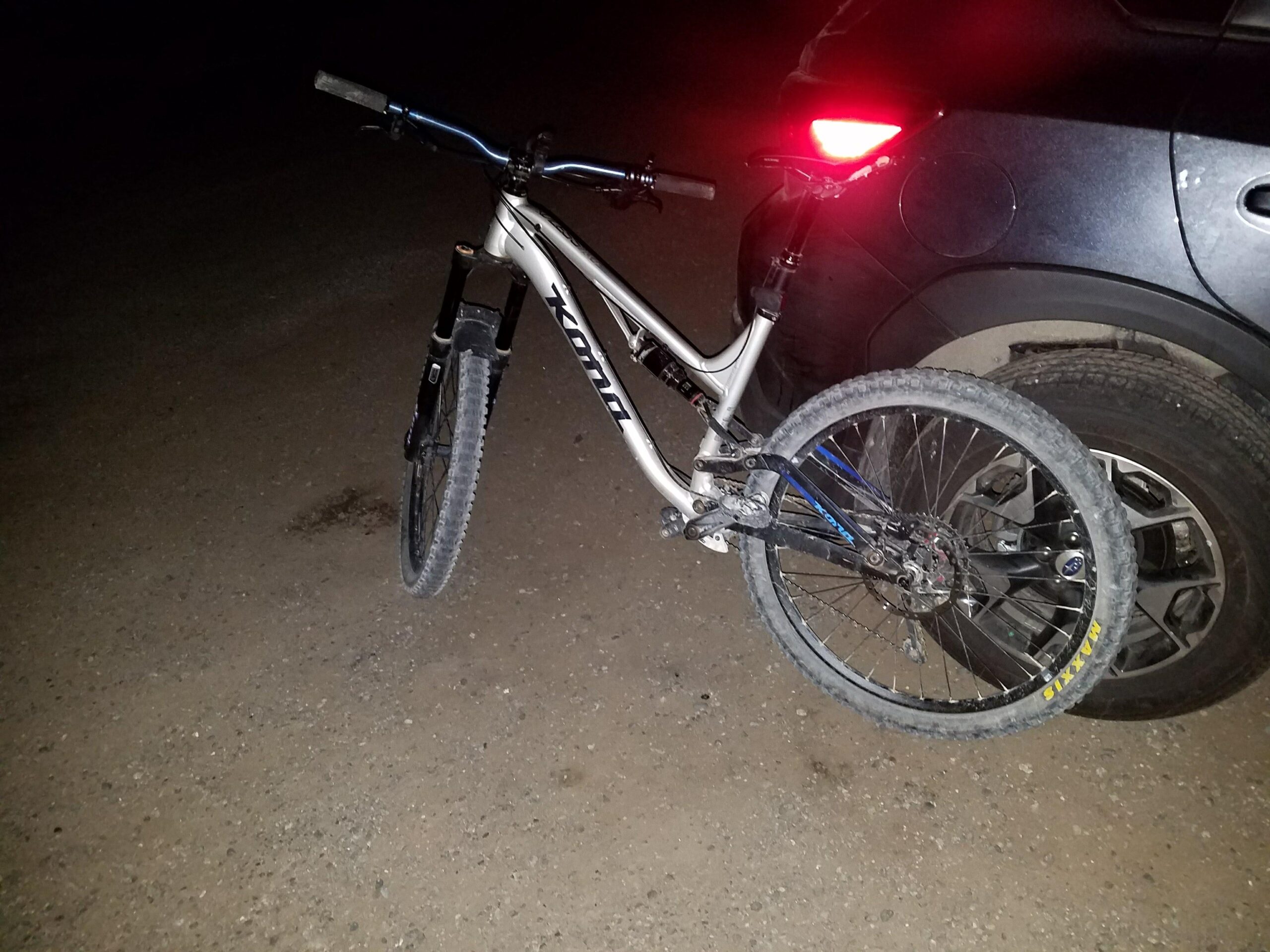 Kona Process 134 DL: A mountain bike with a silver frame is leaning against the rear of a black car at night. The bike's front wheel is suspended above the ground, and the car's tail lights are visible, illuminating the scene. The ground is gravelly and dark.