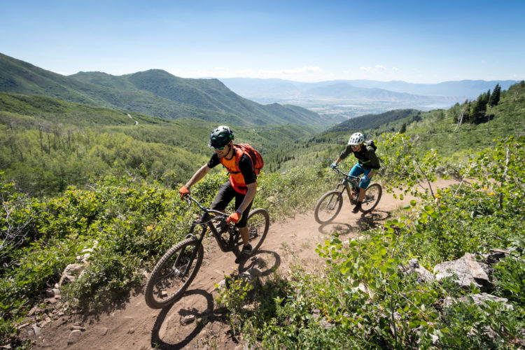 Two mountain bikers riding along a dirt trail through lush green hills under a bright blue sky. The terrain features rolling mountains in the background, and the bikers, wearing helmets and bright clothing, are navigating the winding path.