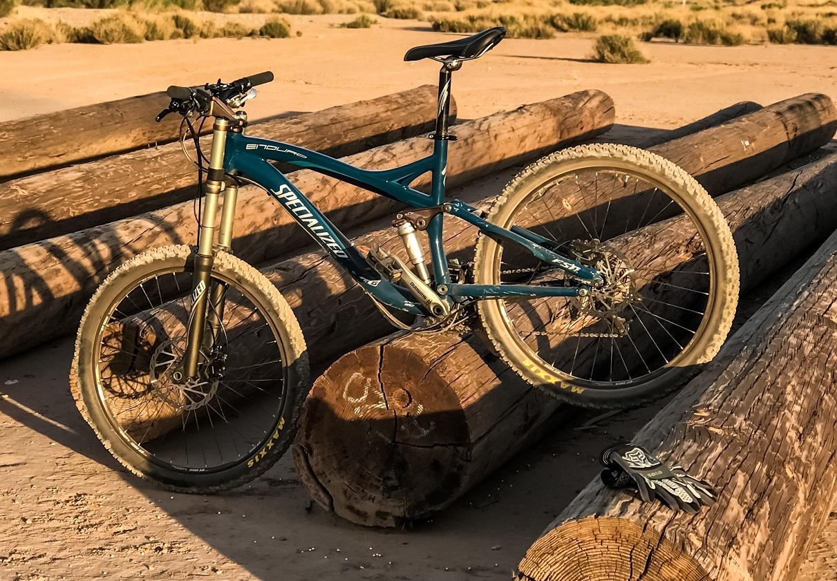 Specialized Enduro: A mountain bike resting on wooden logs in a sandy outdoor setting, with a pair of black gloves nearby. The bike features a teal frame and is partially covered in dirt, indicating it has been used for off-road riding. The background shows dry grass and terrain.