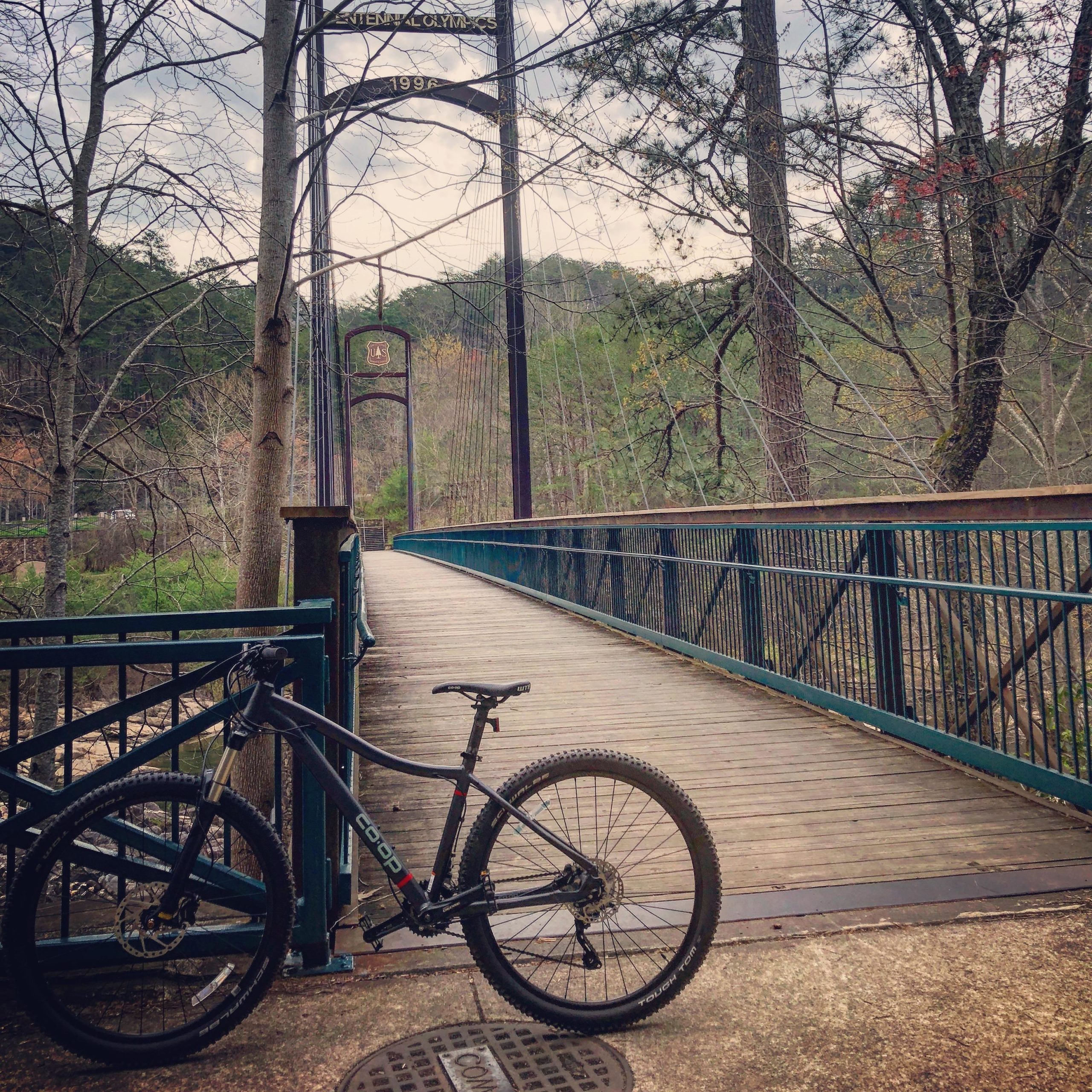 A black mountain bike is parked next to a wooden bridge with a green metal railing. The bridge spans a wooded area, with trees visible in the background and a cloudy sky above. The scene conveys a serene natural setting, ideal for outdoor activities. Tanasi Trail System mountain bike trail.