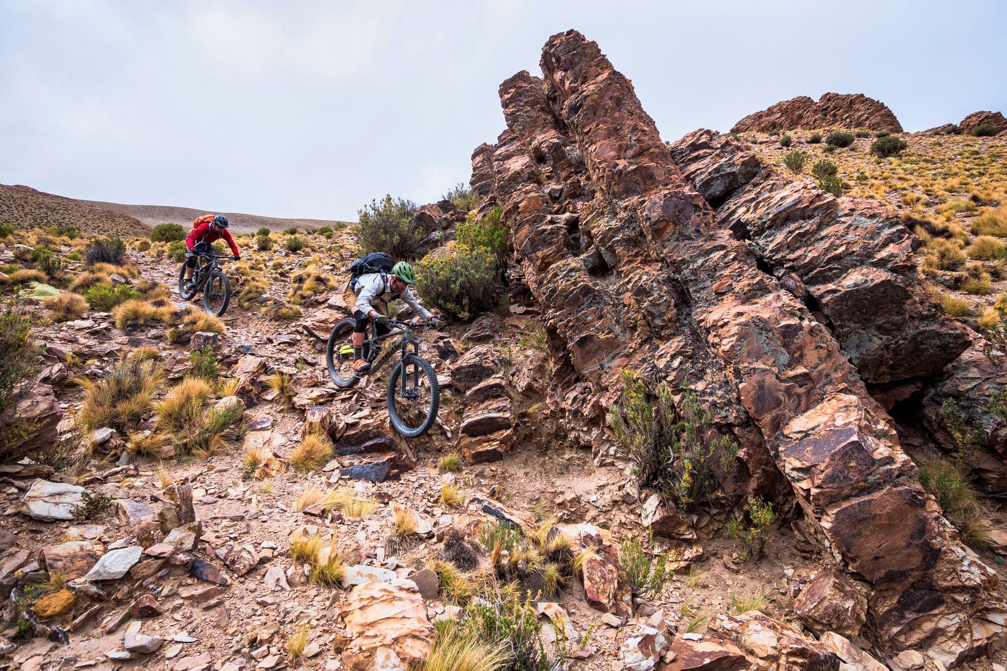 Trek Full Stache: Two mountain bikers navigate a rocky, mountainous terrain with sparse vegetation, under a cloudy sky. One rider, dressed in a red shirt, is climbing uphill, while the other, in a light-colored outfit and green helmet, is focused on descending a steep section of the trail surrounded by rugged rocks.