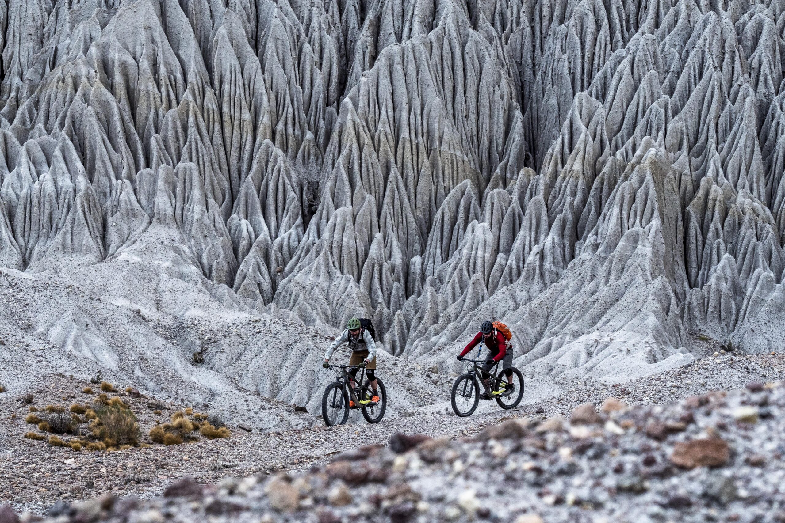 Trek Full Stache: Two mountain bikers navigate a rugged, jagged landscape featuring white and gray formations. The background consists of striking, textured cliffs, while the foreground shows a mix of gravel and sparse vegetation. The scene conveys a sense of adventure in a unique natural setting.