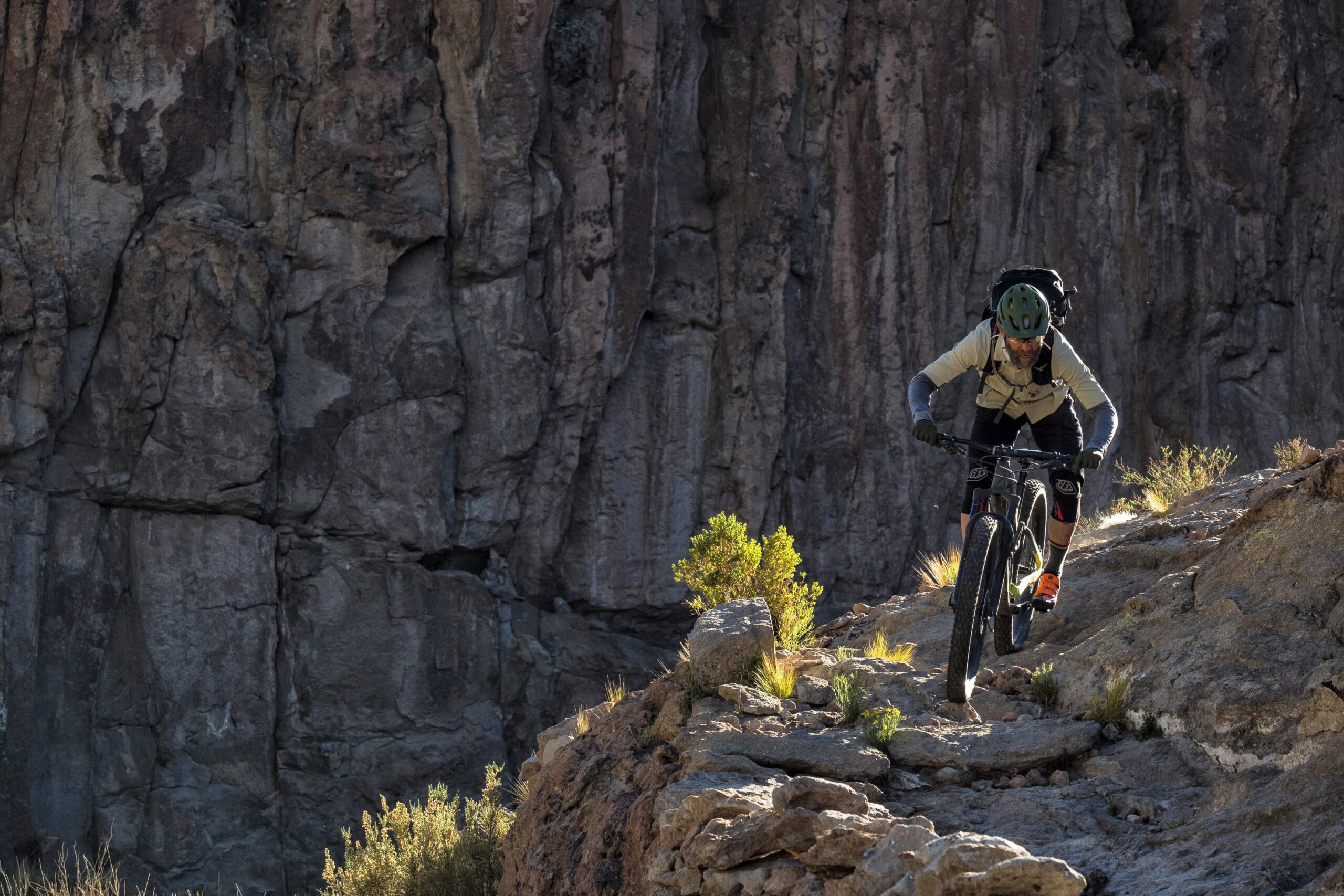Trek Full Stache: A mountain biker navigating a rocky trail on a cliffside, surrounded by rugged terrain and steep rock formations, with the sun casting dramatic shadows. The cyclist is positioned on a narrow path, showcasing a focused riding posture, wearing a helmet and sport gear. Vegetation peeks through the rocky ground, adding a splash of green against the natural backdrop.