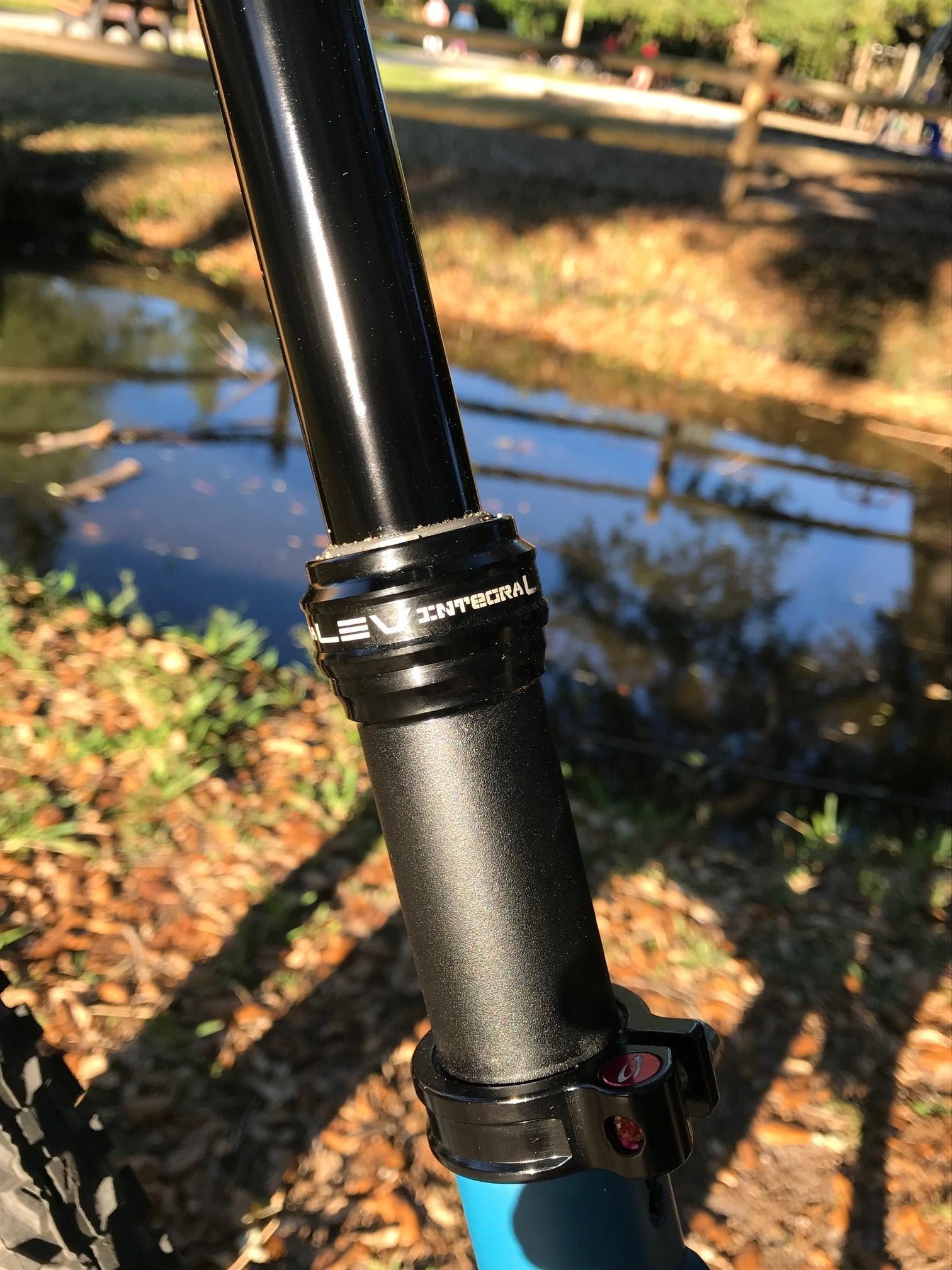 Niner Jet 9: Close-up of a black dropper seat post installed on a mountain bike, featuring a lever for adjustment. The background includes a serene park scene with a pond and green grass. Sunlight casts shadows on the ground, highlighting fallen leaves.