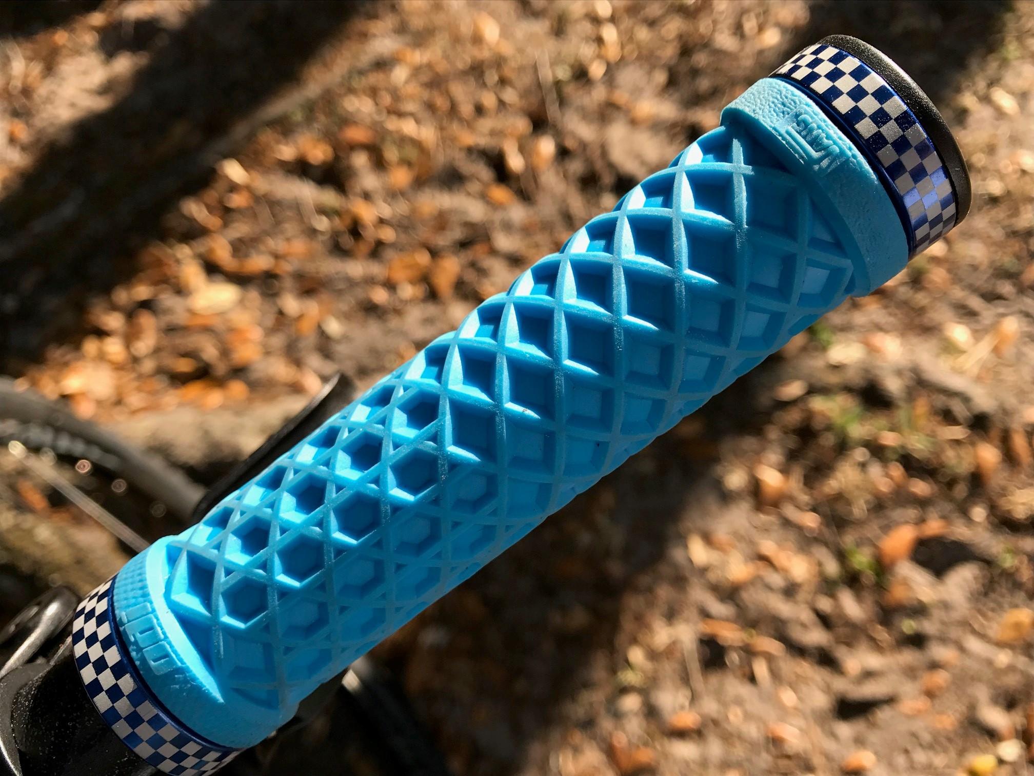 Niner Jet 9: Close-up of a blue bike grip with a textured diamond pattern, featuring a checkered design at the end. The background shows blurred natural elements, like dirt and fallen leaves, indicating an outdoor setting.