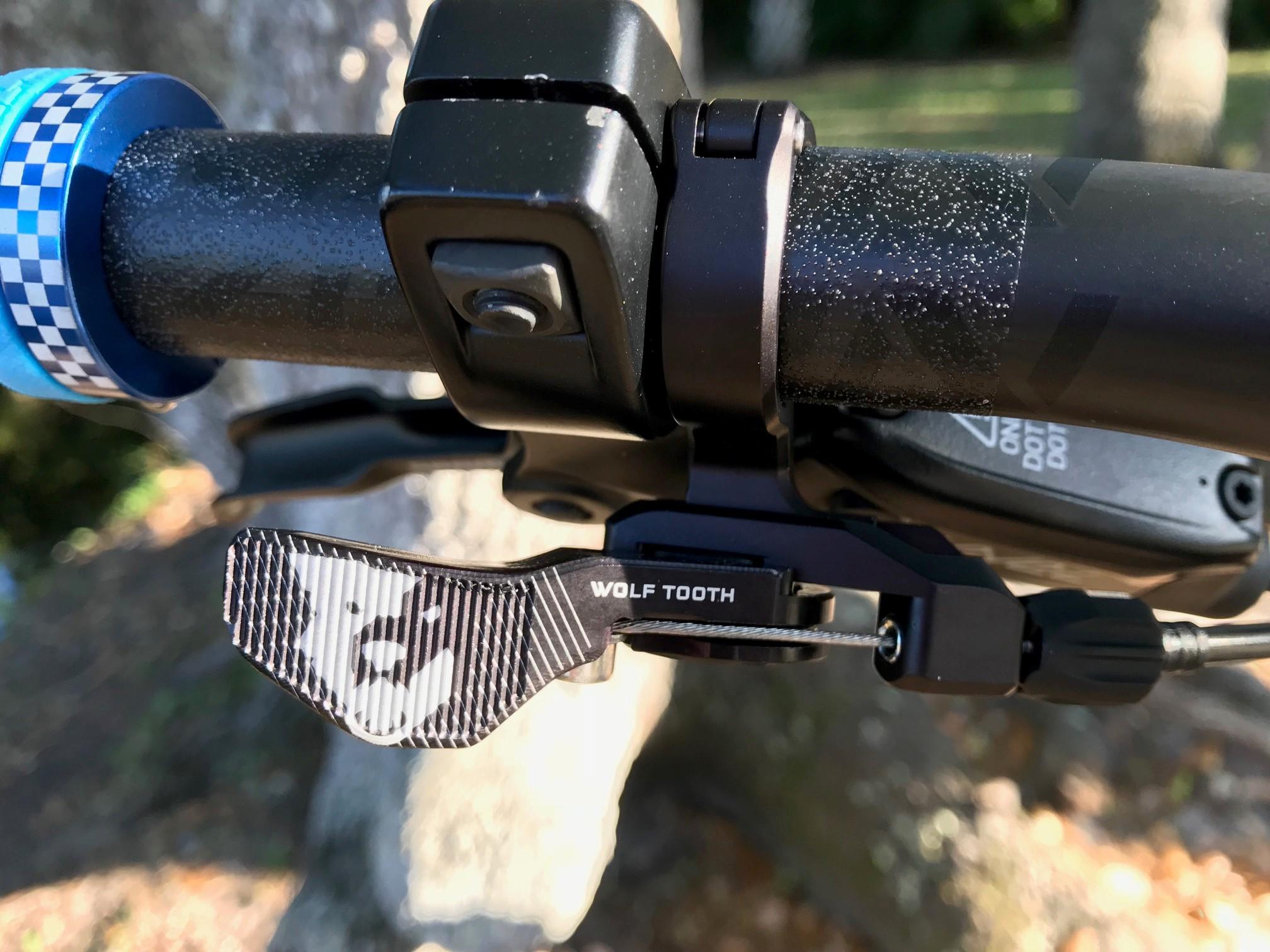 Niner Jet 9: Close-up of a bicycle handlebar showing a Wolf Tooth dropper post remote lever, featuring a textured black and white design. The handlebar grips have a blue and white checkered pattern, indicating a sporty design. Natural background with dappled light from surrounding trees.