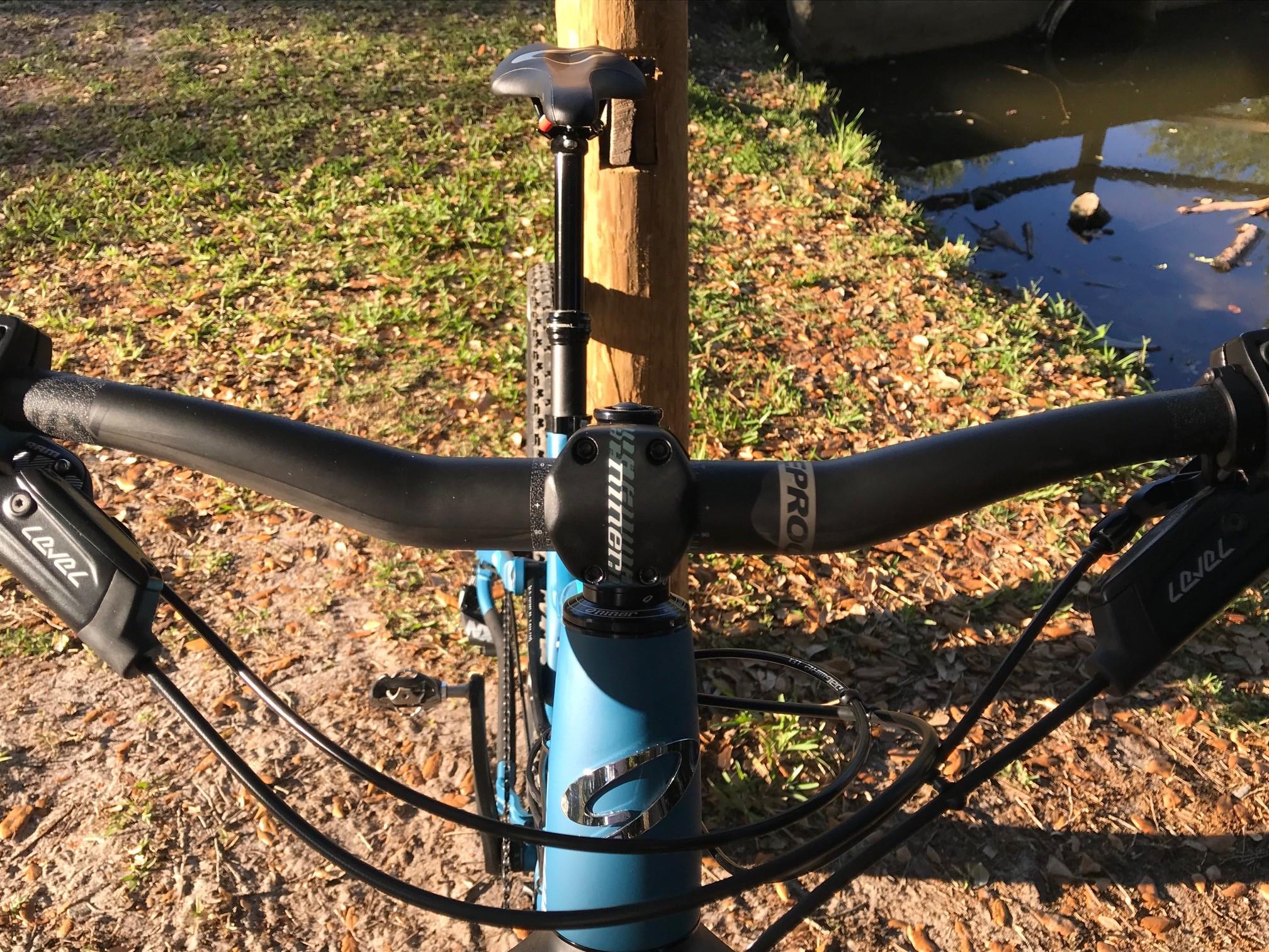 Niner Jet 9: A close-up view of a mountain bike's handlebars, showcasing the grips, shifters, and cables. The bike is positioned near a wooden post with a natural setting in the background, including grass and a pond. The sunlight casts a warm glow on the scene, highlighting the bike's blue frame and components.