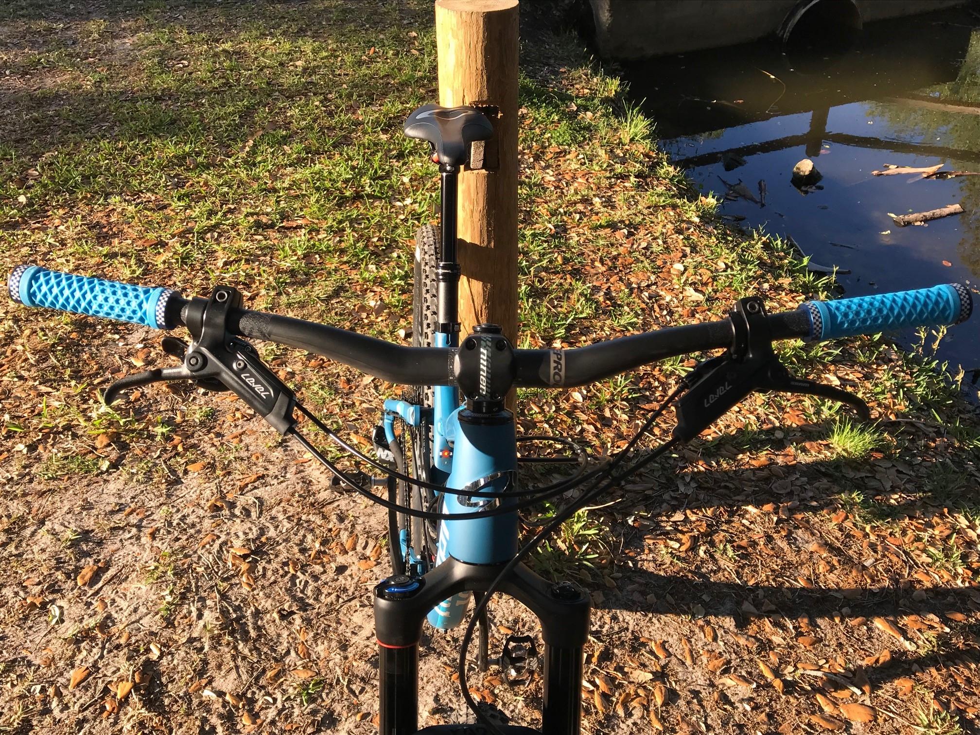 Niner Jet 9: Close-up view of a mountain bike's handlebars, featuring blue grips, brake levers, and a gear shifter. The bike is positioned outdoors on grassy ground near a body of water, surrounded by fallen leaves.