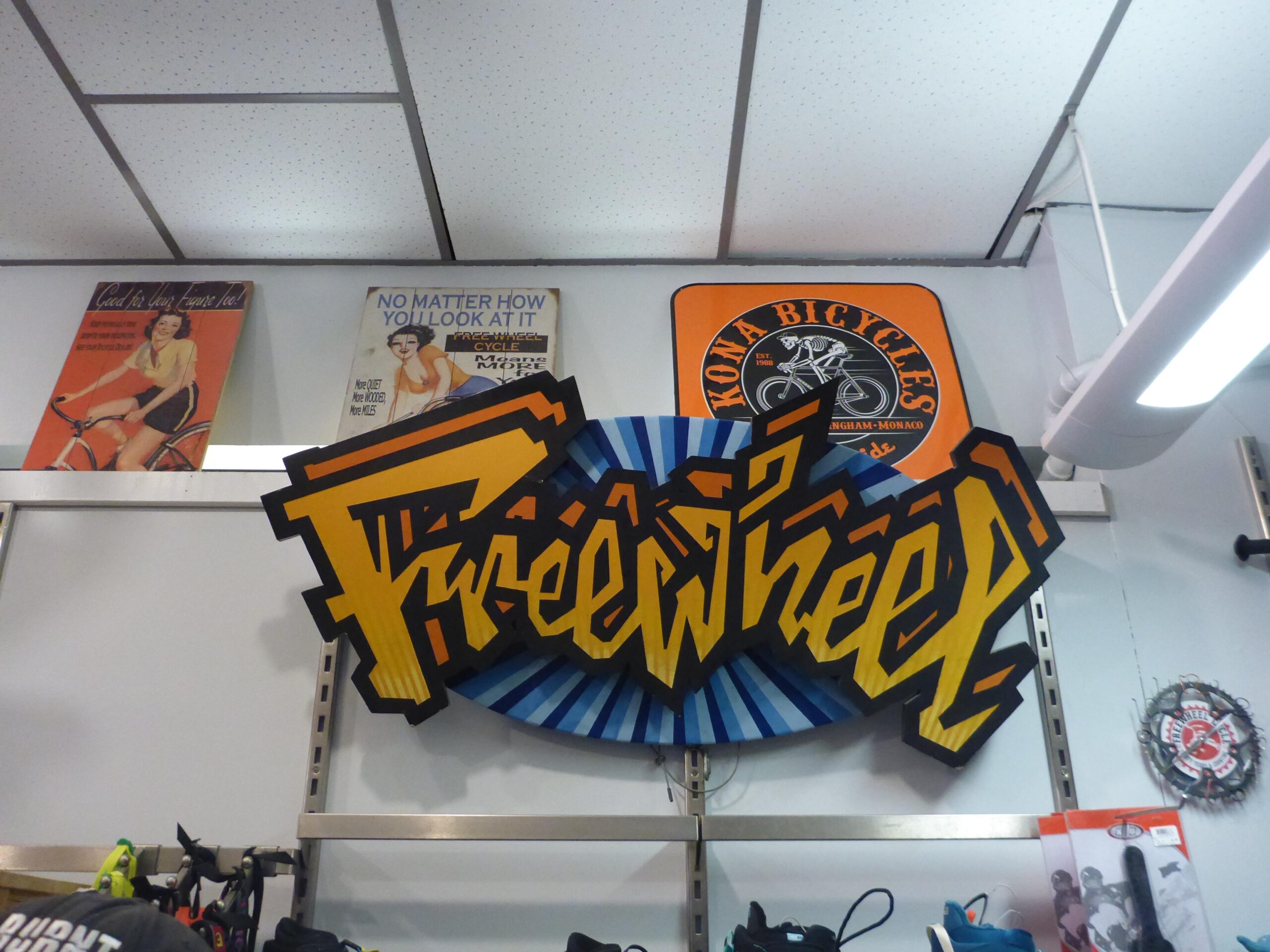 Colorful sign reading "Freewheel" displayed on a wall, with vintage cycling-themed posters above and a Kona Bicycles sign nearby. Various cycling gear can be seen on a shelf below.