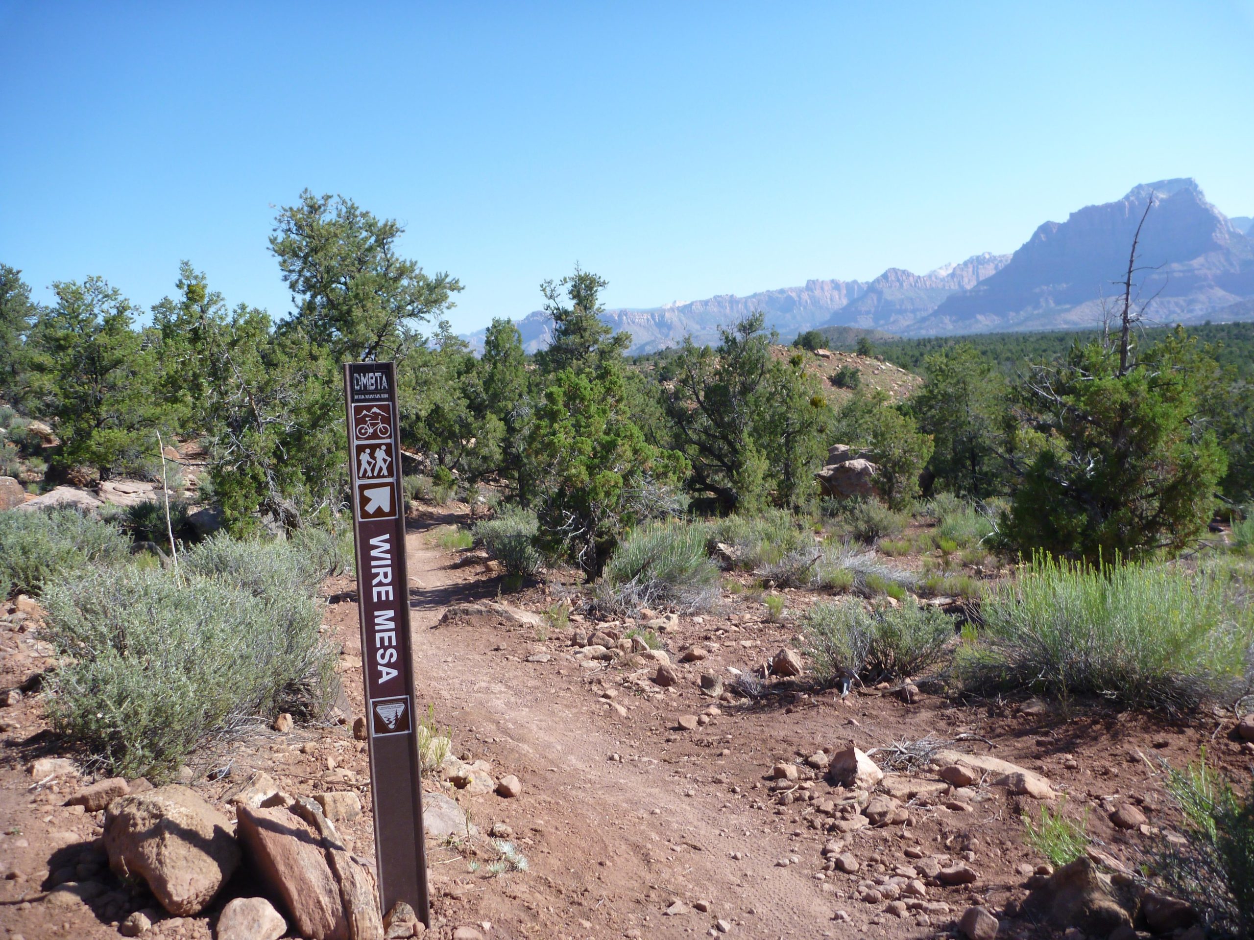 Trail sign for Wire Mesa indicating directions for hikers and bikers, with a scenic view of mountains and dense greenery in the background on a clear day. Wire Mesa Loop mountain bike trail.