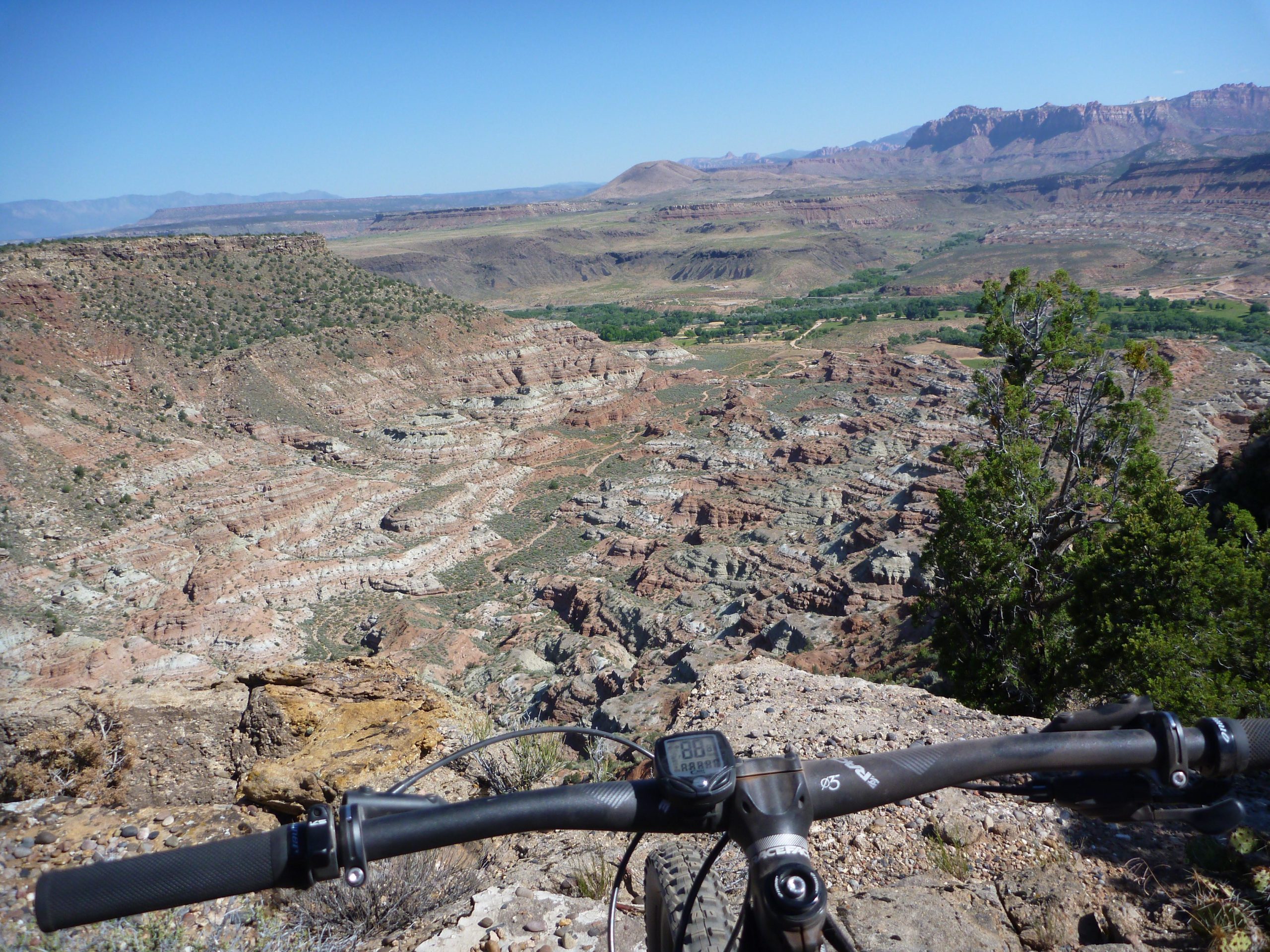 Mountain bike handlebars in the foreground with a panoramic view of a rugged canyon landscape featuring layered rock formations, greenery, and distant mountains under a clear blue sky. Wire Mesa Loop mountain bike trail.