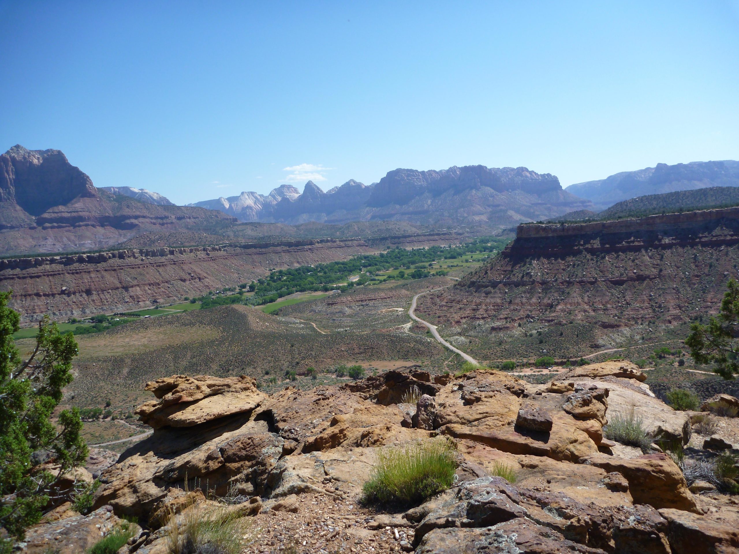 A panoramic view of a rugged landscape showcasing layered rock formations, rolling hills, and a lush green valley. In the foreground, large rocky outcrops are visible, while distant mountains rise against a clear blue sky, suggesting a serene natural environment. A winding road can be seen running through the valley. Wire Mesa Loop mountain bike trail.