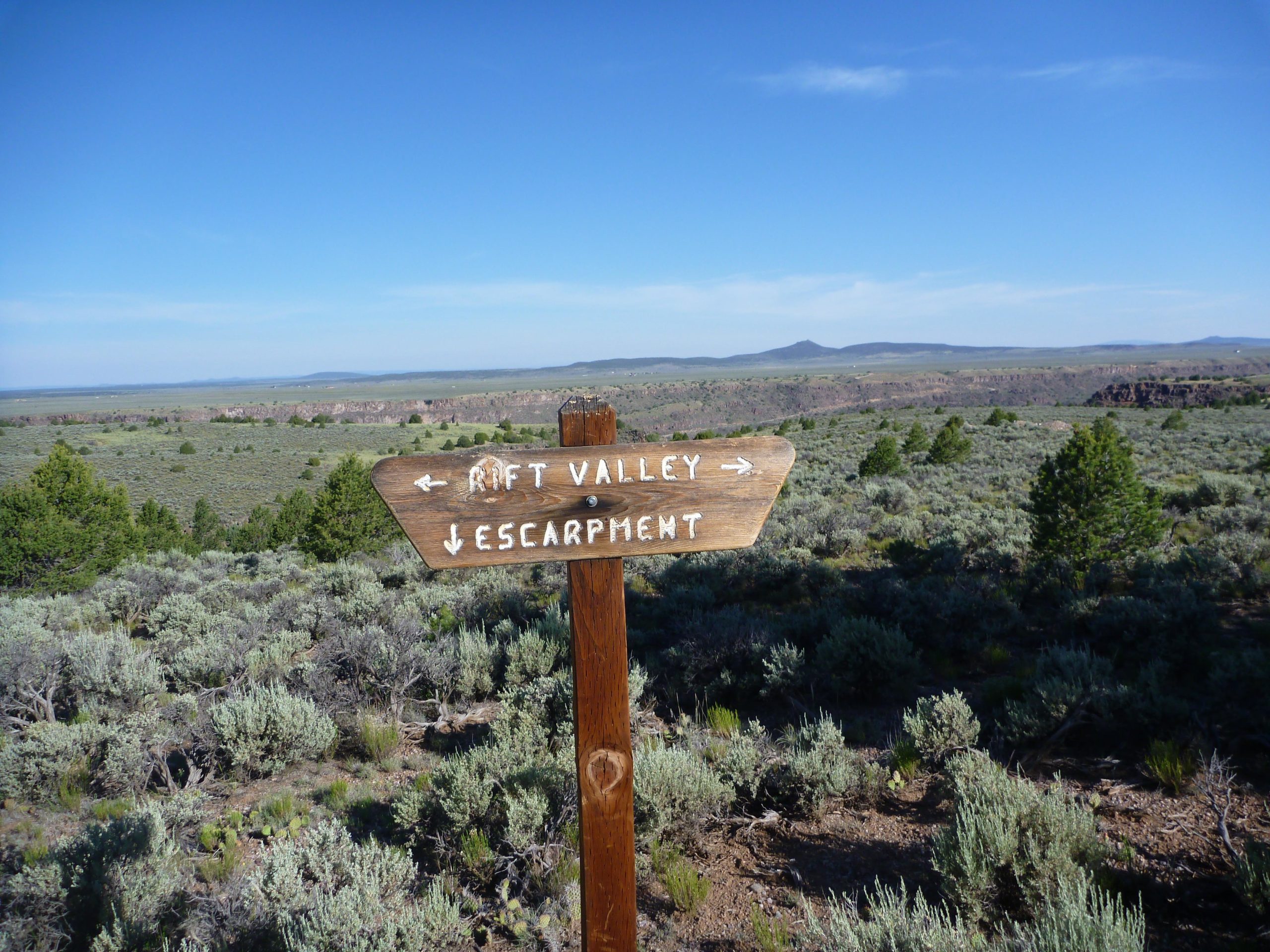 Signpost indicating directions to "Rift Valley" and "Escarpment" in a scenic landscape characterized by sagebrush and distant mountains under a clear blue sky. Taos Valley Overlook mountain bike trail.