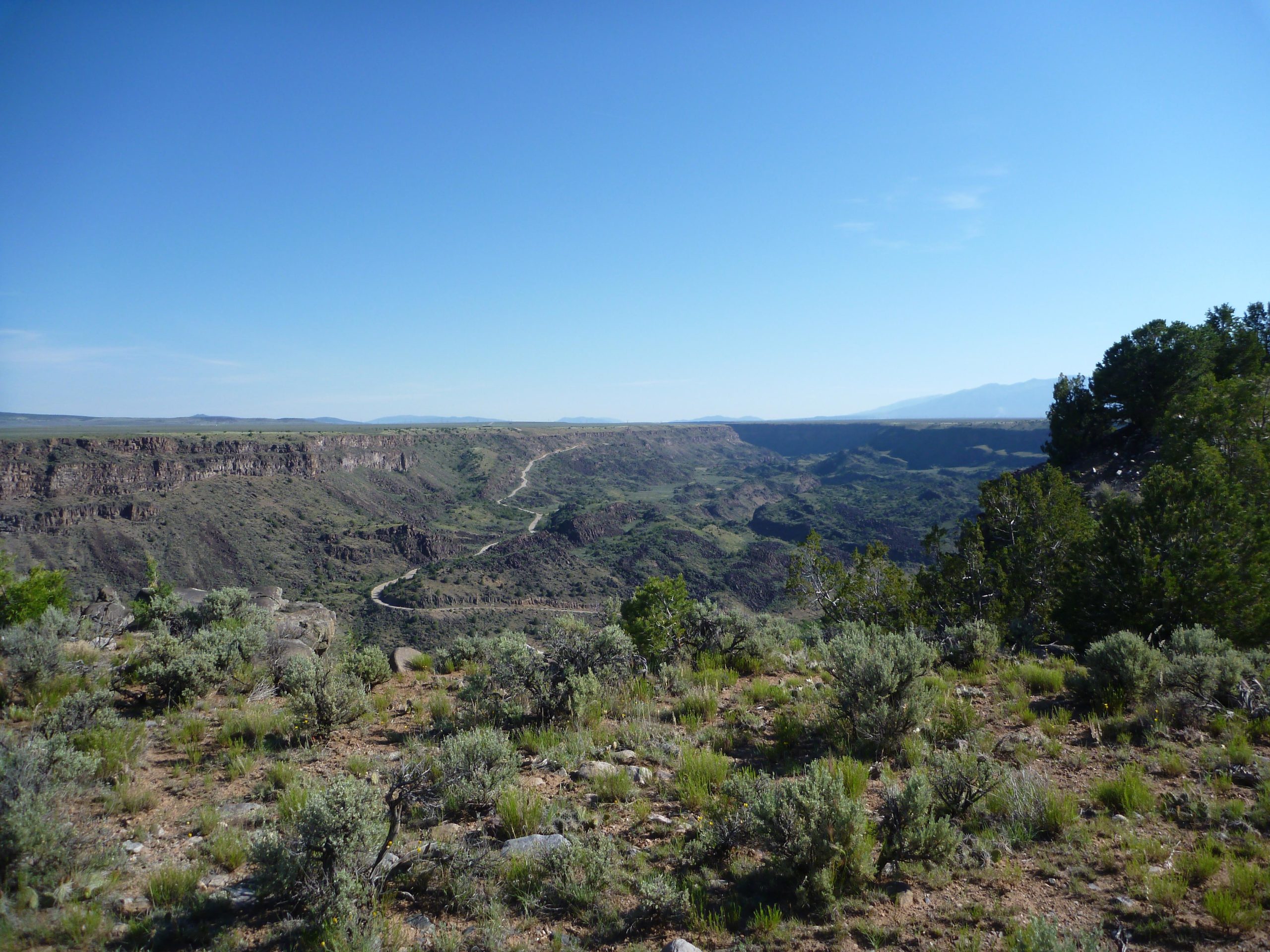 A panoramic view of a canyon landscape under a clear blue sky, featuring rocky cliffs and a winding road. The foreground showcases sparse vegetation, including shrubs and grasses, while the expansive background reveals rolling hills and distant mountains. Taos Valley Overlook mountain bike trail.