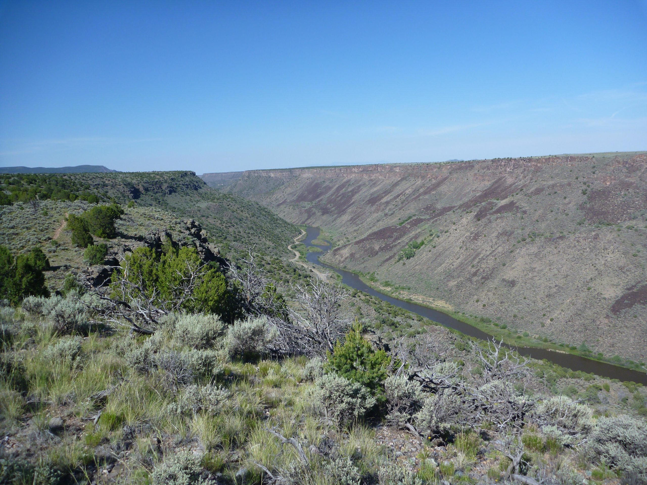 A scenic view of a winding river flowing through a rugged canyon landscape. The foreground features shrubbery and sparse vegetation, while the canyon walls rise steeply on either side, displaying layers of earth. The sky above is clear and blue, creating a bright, sunny atmosphere. Taos Valley Overlook mountain bike trail.