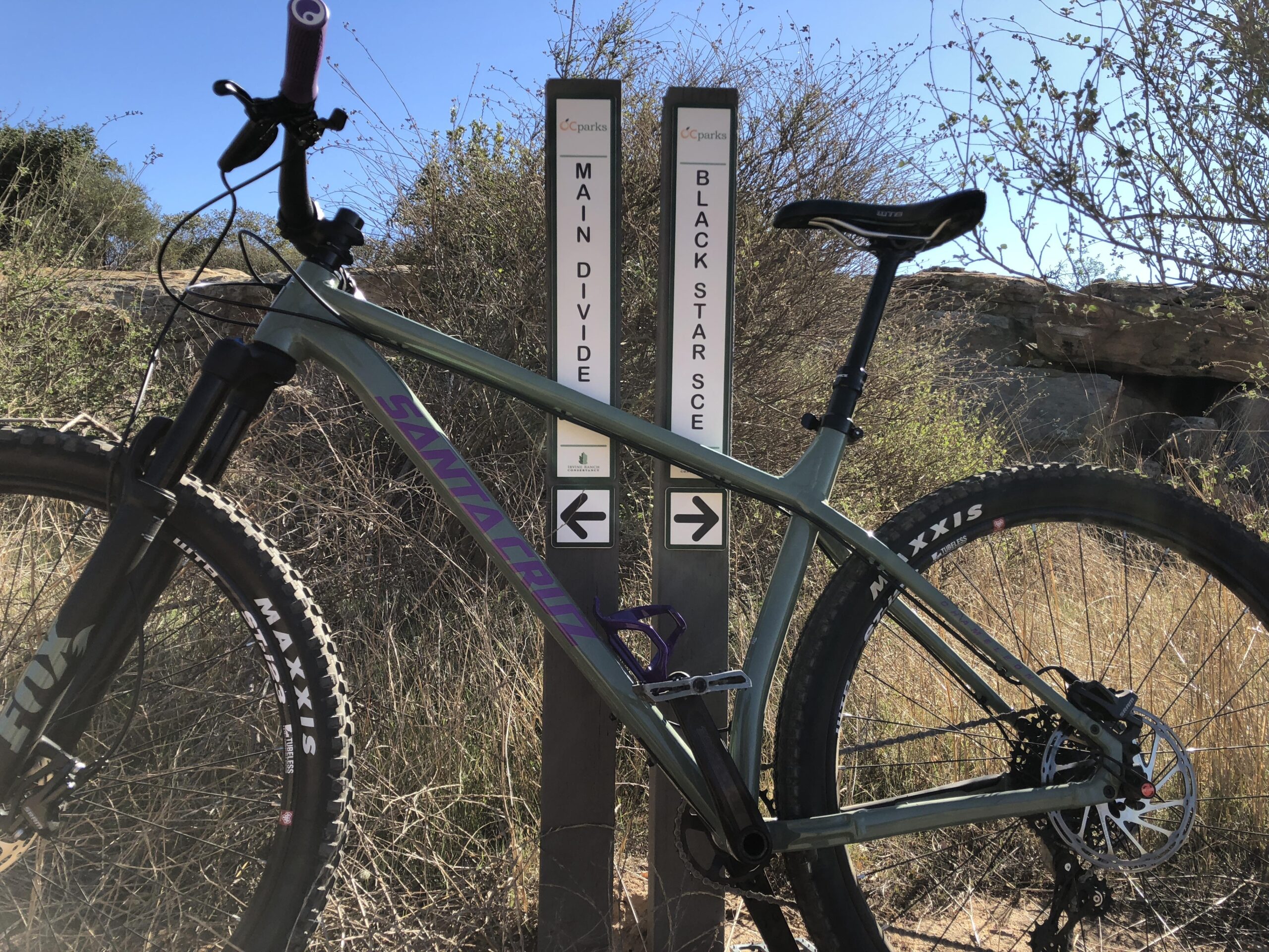 Santa Cruz Chameleon: A mountain bike is parked next to two directional signs for hiking and biking trails labeled "Main Divide" and "Black Star SCE," set against a backdrop of dry brush and clear blue skies.