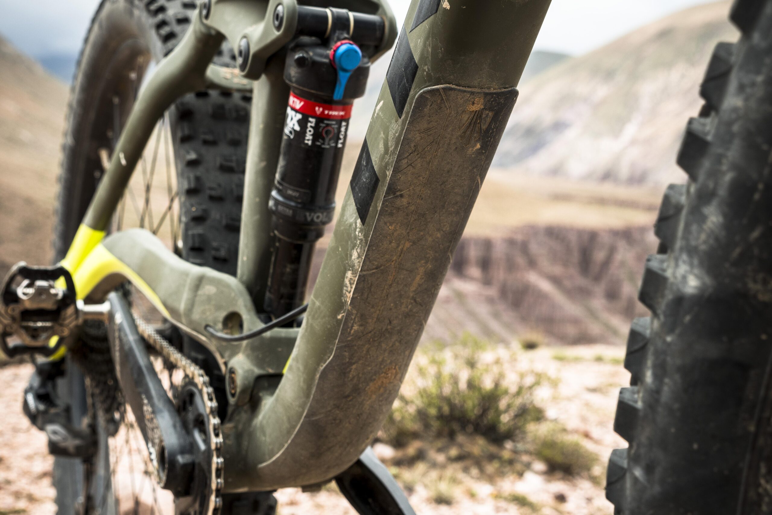 Trek Full Stache: Close-up view of a mountain bike frame featuring a slightly muddy green and yellow design, highlighting the rear suspension and chain mechanisms against a rugged outdoor backdrop.