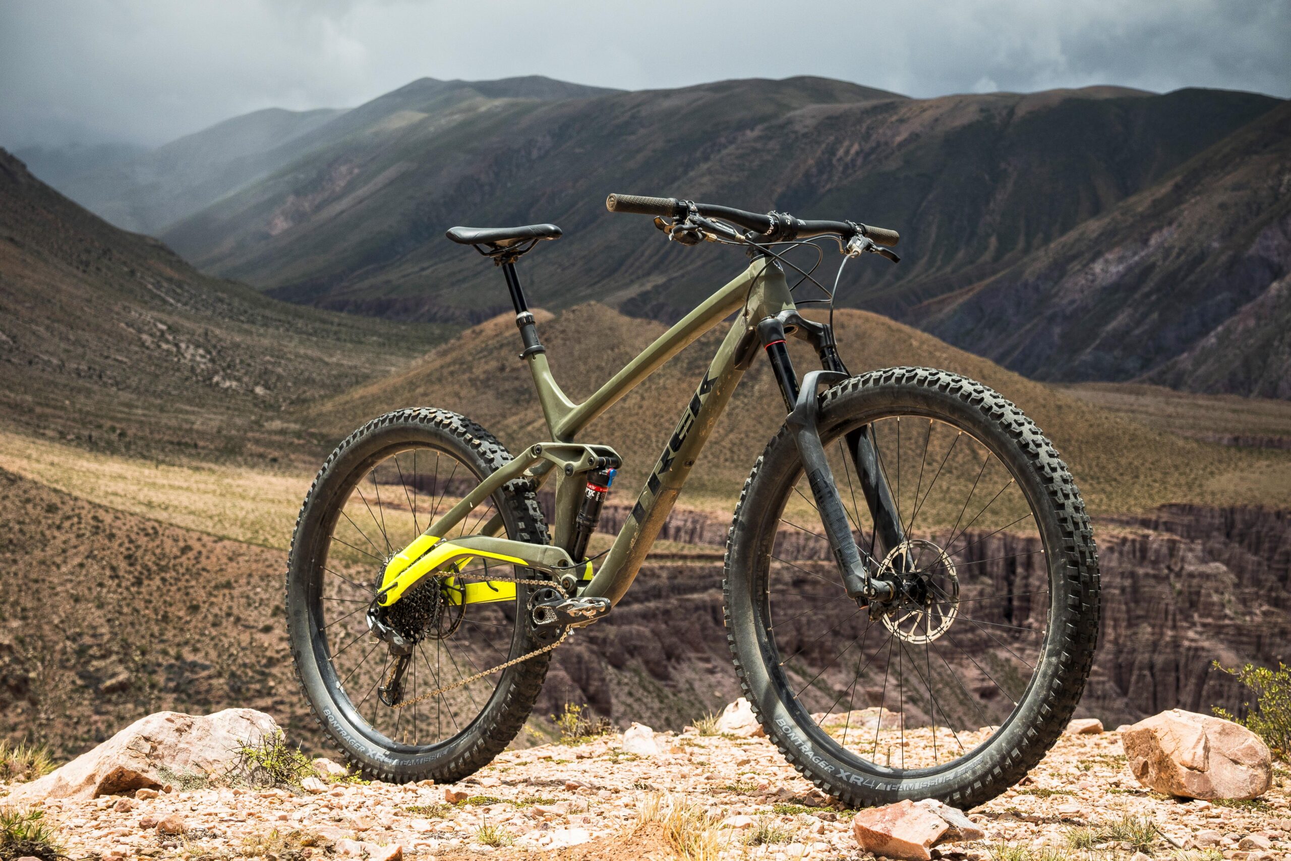 Trek Full Stache: A mountain bike positioned on rocky terrain with a scenic backdrop of rugged mountains and a cloudy sky. The bike features a lightweight frame, wide tires with tread patterns suitable for off-road riding, and a bright yellow detailing on the rear. The landscape shows undulating hills and valleys, creating a dramatic outdoor setting.