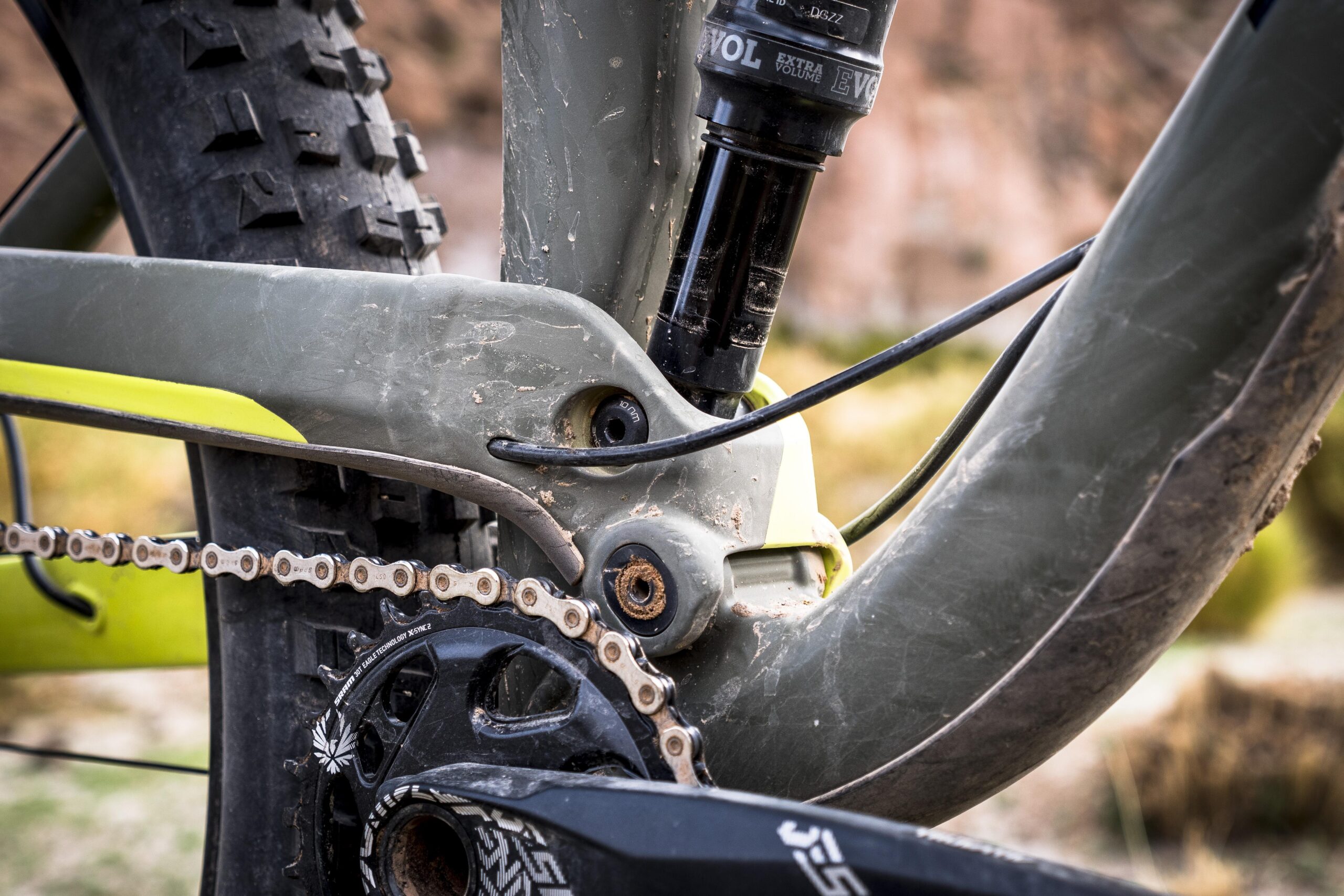 Trek Full Stache: Close-up of a mountain bike's bottom bracket area, showcasing the chain, crankset, suspension components, and tire. The bike frame is green with yellow accents, and it appears slightly dirty, indicating usage on rugged terrain.