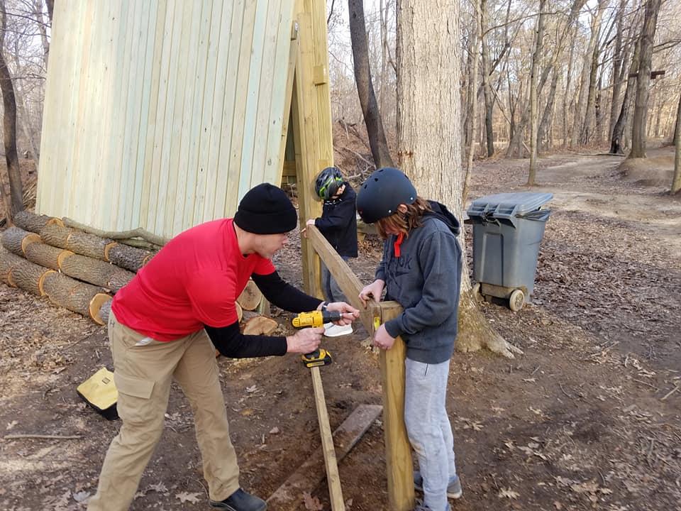 A man and two children are working together outdoors to build a wooden structure. The man, wearing a red shirt and a black beanie, is using a power drill to secure a wooden post while guiding one of the children. The other child, wearing a helmet, observes closely in the background. A tree and a partially constructed wooden structure can be seen, along with some logs on the ground and a trash bin nearby, indicating a natural setting. Scales Lake Park Trails mountain bike trail.