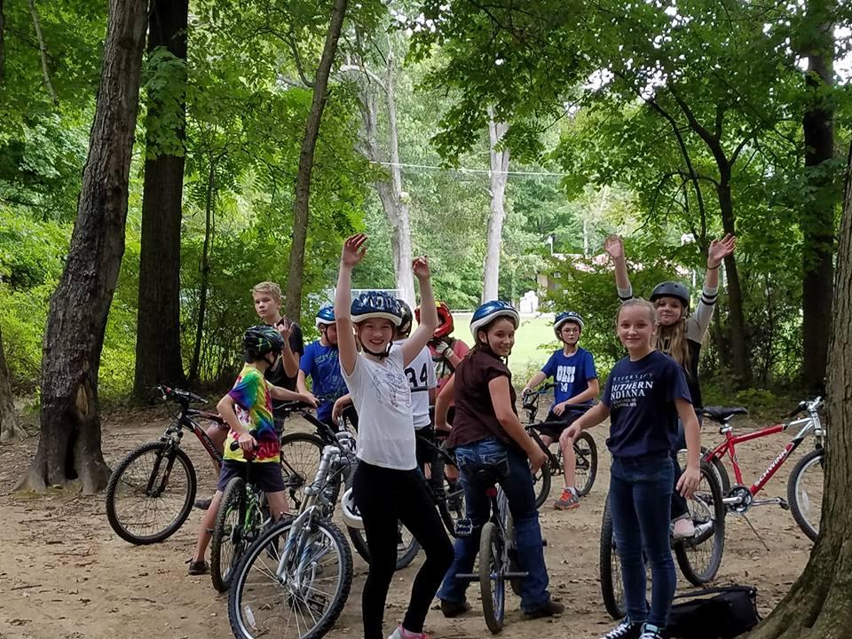 A group of children enjoying a fun day of mountain biking in a wooded area. They are wearing helmets and smiling, with some of them standing next to their bikes while others are seated. The scene conveys excitement and camaraderie in an outdoor setting surrounded by lush greenery. Scales Lake Park Trails mountain bike trail.