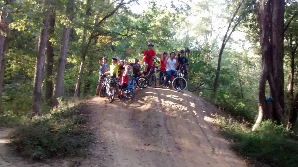 A group of children and teens with bicycles are posing on a dirt ramp in a wooded area. They are wearing helmets and colorful clothing, surrounded by tall trees and greenery. The scene captures a fun, active moment outdoors. Scales Lake Park Trails mountain bike trail.