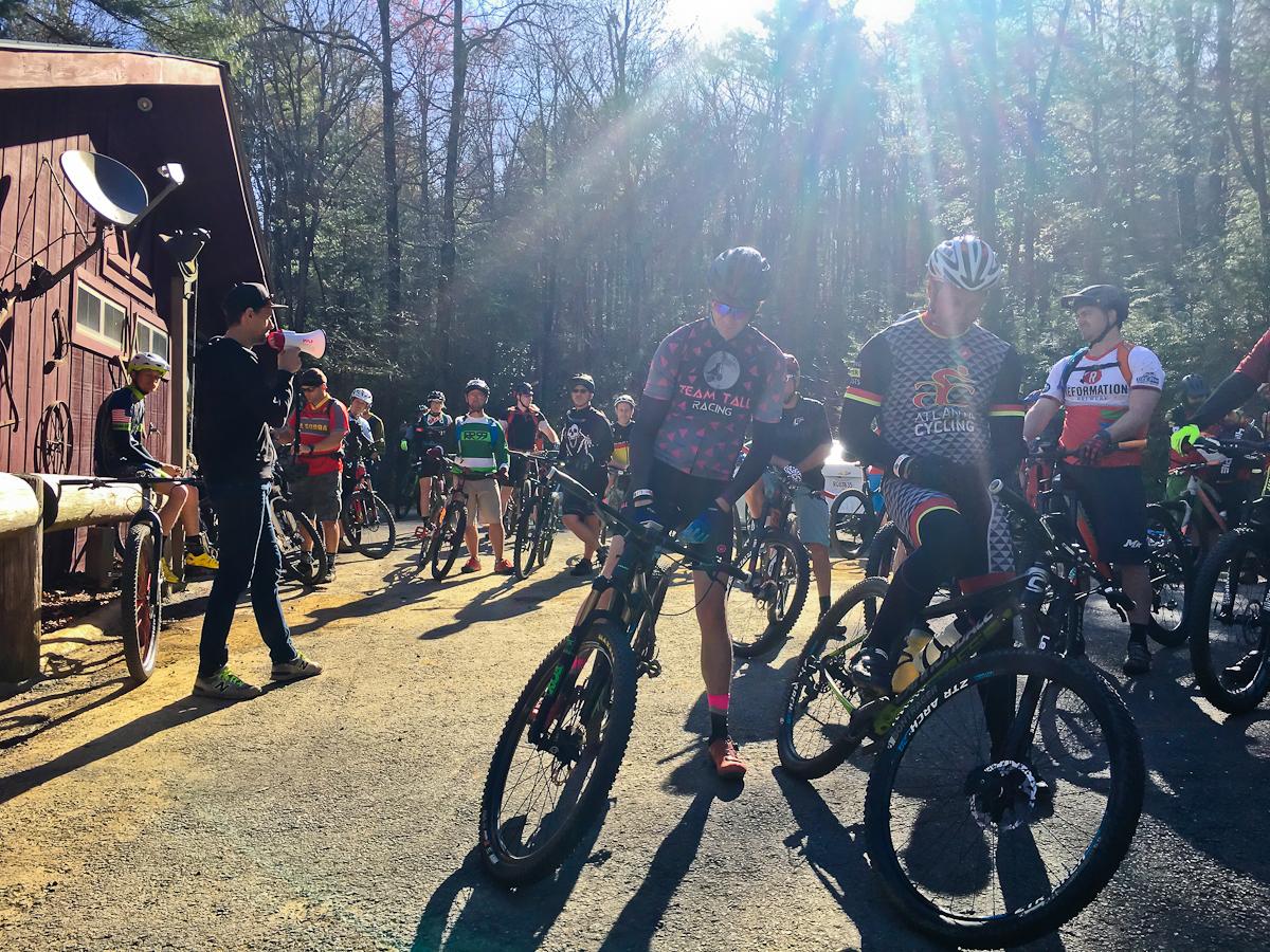 A group of mountain bikers gathered outside a rustic building, listening to a speaker using a megaphone. The scene is set in a wooded area under bright sunlight, with various cyclists wearing colorful jerseys and helmets, preparing for a ride. Bikes are parked around them on a dirt path. Pinhoti Trail: Mountaintown Creek Segment mountain bike trail.