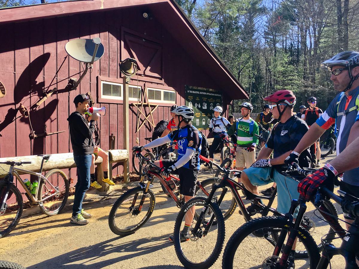 A group of mountain bikers gathered outside a wooden building, listening to a speaker using a megaphone. The scene features several cyclists in various riding gear and helmets, with their bikes parked nearby. The background includes a mix of trees and rustic decor on the building, suggesting an outdoor event or gathering. Pinhoti Trail: Mountaintown Creek Segment mountain bike trail.