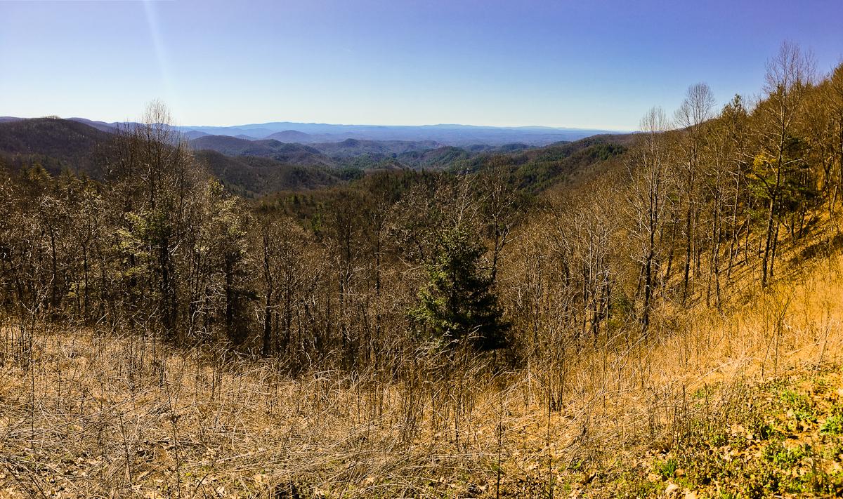 A panoramic view of rolling mountains under a clear blue sky, featuring a landscape with sparse trees and dry grass in the foreground. The backdrop showcases layers of distant hills, creating a serene natural scene. Pinhoti Trail: Mountaintown Creek Segment mountain bike trail.