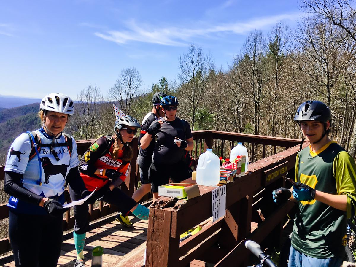 A group of mountain bikers gathered on a wooden deck in a forested area, enjoying a break. The scene features five individuals, some wearing helmets and cycling gear, as well as a picnic table with supplies like water jugs and snacks. The landscape in the background shows rolling hills under a clear blue sky. One member is holding a map while another stretches. A sign indicating the "Brutal Loop" is visible nearby. Pinhoti Trail: Mountaintown Creek Segment mountain bike trail.
