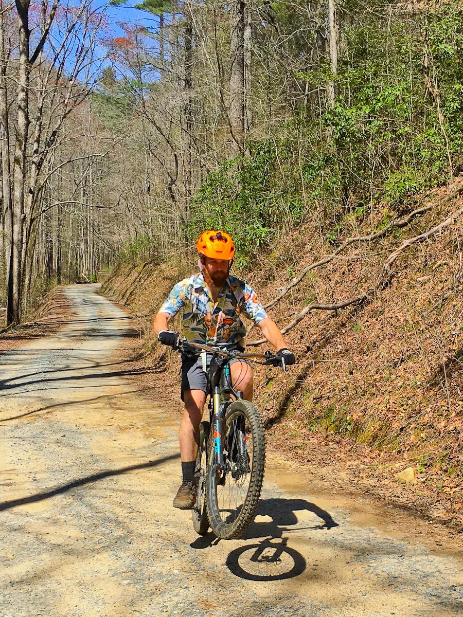 A person riding a mountain bike on a gravel path surrounded by trees, performing a wheelie. The rider is wearing an orange helmet, a colorful shirt, and shorts. Sunlight filters through the trees, illuminating the scene. Pinhoti Trail: Mountaintown Creek Segment mountain bike trail.