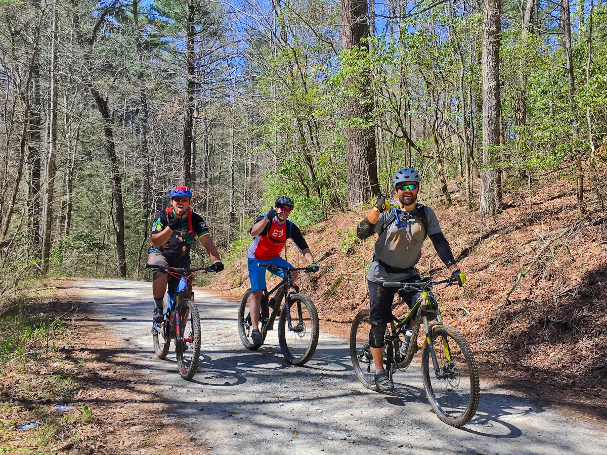 Three mountain bikers are riding along a dirt trail surrounded by trees. Two riders are smiling and giving a thumbs-up, while the third is also smiling and gesturing positively. The scene captures a sunny day with green foliage peeking through the forest. Pinhoti Trail: Mountaintown Creek Segment mountain bike trail.