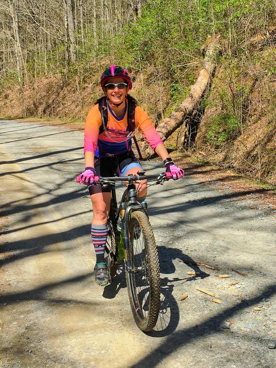 A woman smiles broadly while riding a mountain bike on a gravel path. She is wearing a colorful cycling jersey, a helmet with pink accents, and sunglasses. Her outfit is complemented by vibrant striped knee socks. The background features a wooded area with leafless trees and greenery, suggesting an outdoor setting on a sunny day. Pinhoti Trail: Mountaintown Creek Segment mountain bike trail.