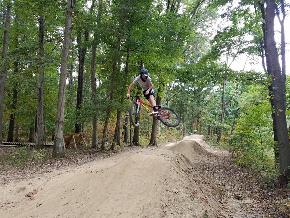 A mountain biker performing a jump over a dirt mound in a wooded area, surrounded by trees with green leaves. The cyclist is wearing a helmet and a gray shirt, showcasing an action-packed moment in a natural setting. Scales Lake Park Trails mountain bike trail.