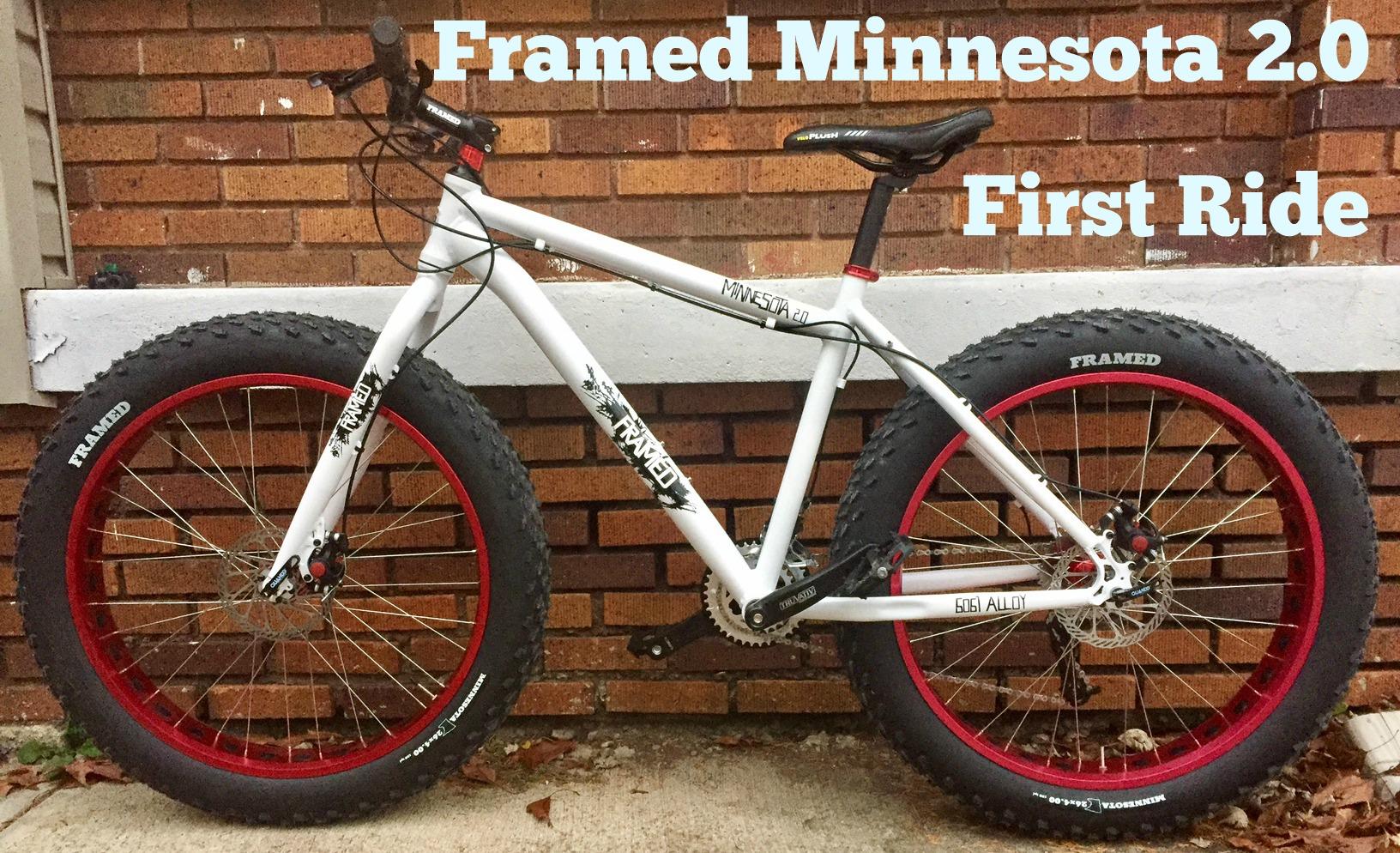 Framed Minnesota 2.0: A white Framed Minnesota 2.0 fat bike with red rims is leaning against a brick wall. The bike features wide tires suitable for various terrains and is set up for its first ride. The background shows a flat, concrete surface with some fallen leaves.