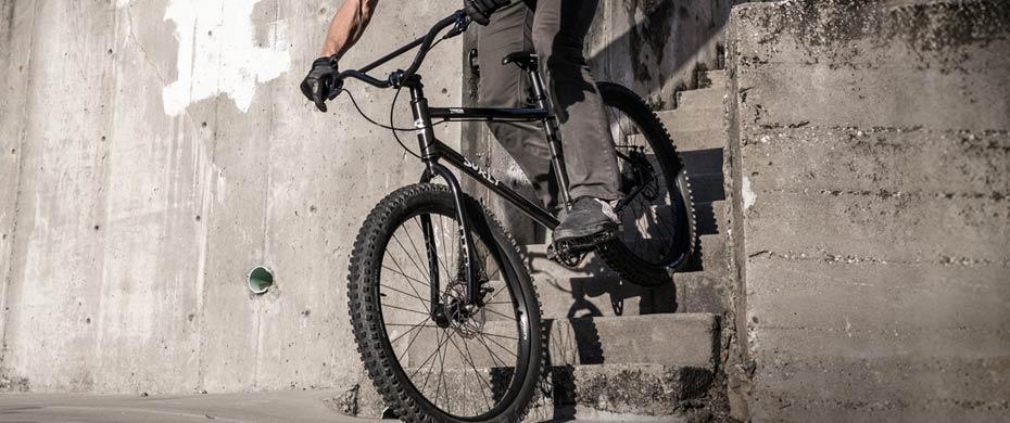 Surly Lowside: A cyclist riding a black mountain bike down a set of concrete stairs, demonstrating skills in an urban environment. The scene captures the dynamic movement and the texture of the surrounding concrete.