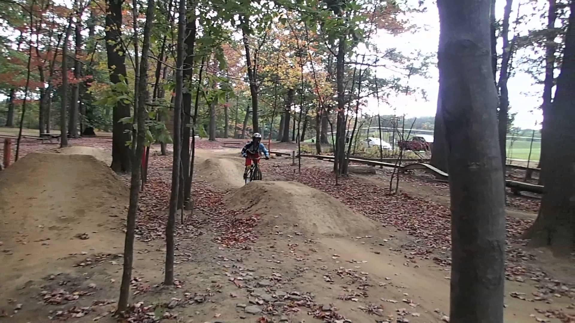 A person riding a bicycle over a dirt jump in a wooded area during autumn. The ground is covered with fallen leaves, and trees surround the bike path, creating a natural setting. The rider is wearing protective gear, including a helmet, and appears to be focused on navigating the jump. North Park mountain bike trail.