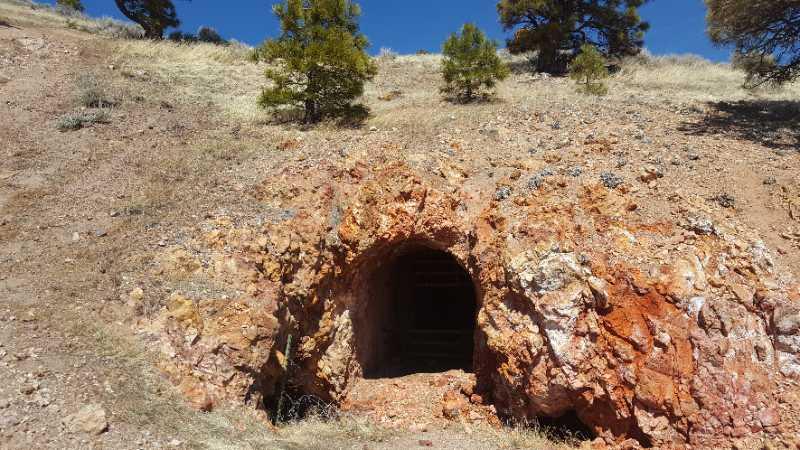 A weathered rock entrance to a mine or cave, surrounded by dry, barren land with sparse vegetation. The entrance reveals a dark interior, suggesting depth, while the rocky textures feature shades of orange and brown under a clear blue sky. Peavine mountain bike trail.