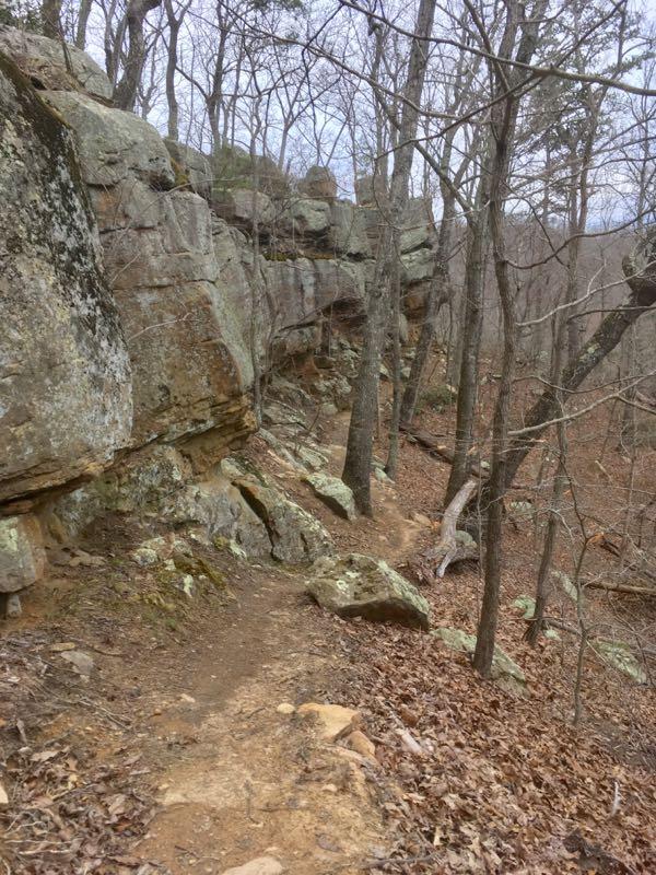 A rocky trail winding through a forest with bare trees and fallen leaves on the ground. Large boulders and a rock face form a rugged backdrop along the path. Sugarcamp Mountain mountain bike trail.